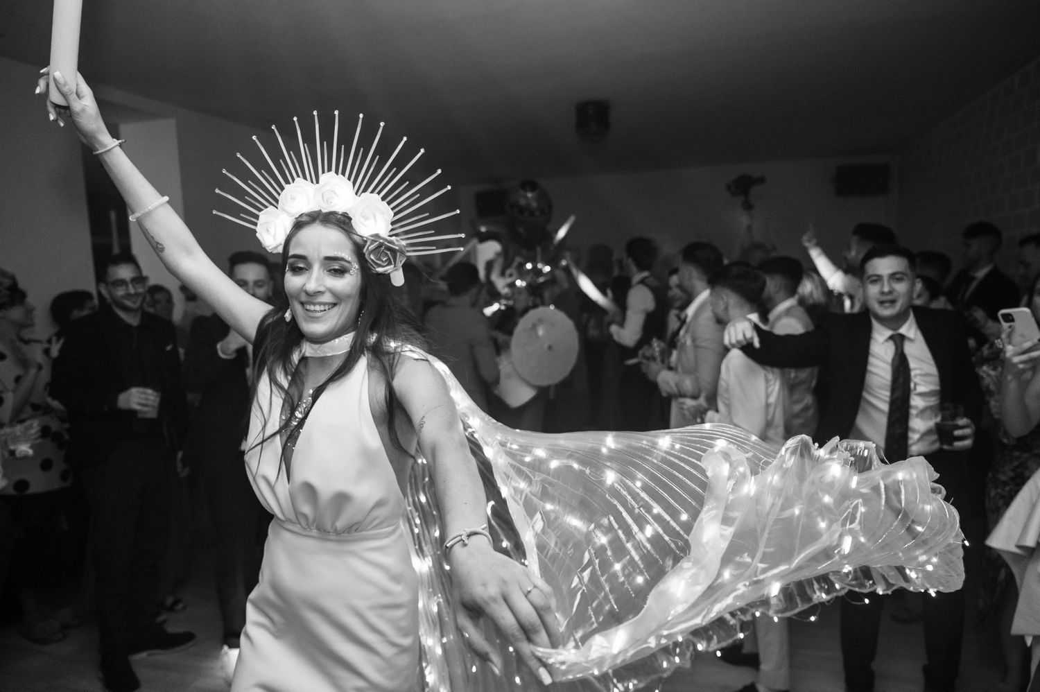 A black and white photo of a dancing woman in white dress and crown with arms raised in celebration.