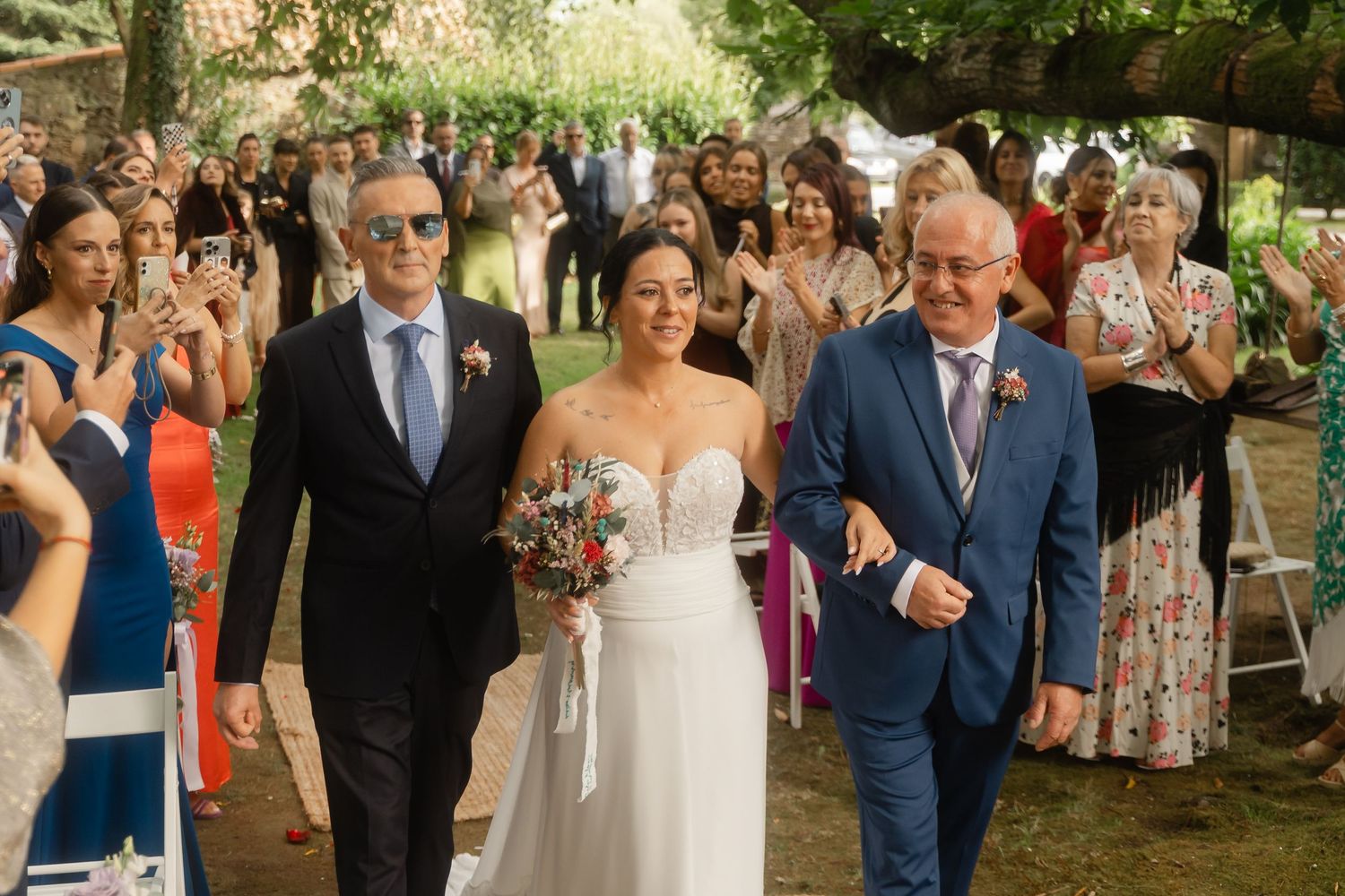 A bride in a white strapless gown walks down an outdoor garden path while guests look on during a summer wedding ceremony.