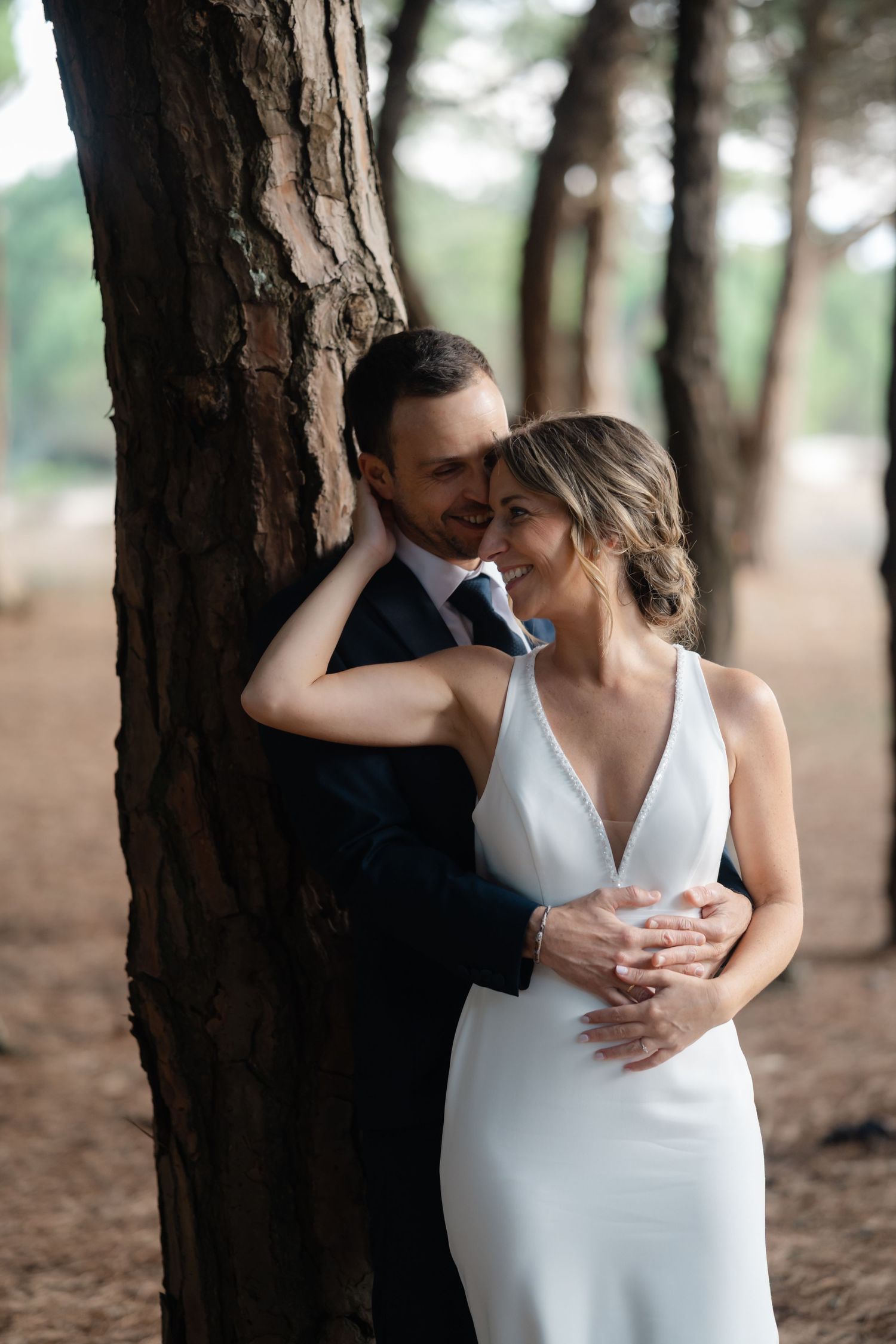 Romantic wedding couple embraces against a rustic tree trunk in a pine forest setting.