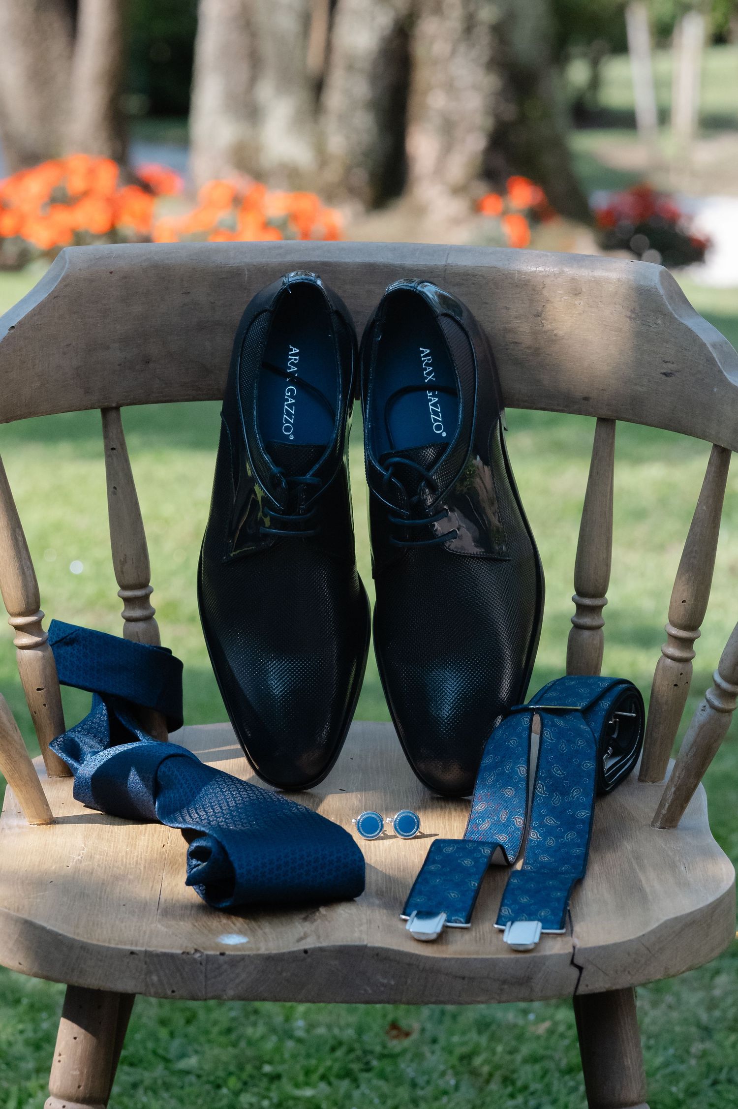 Elegant black dress shoes and navy blue accessories arranged on a wooden chair with orange flowers in background.