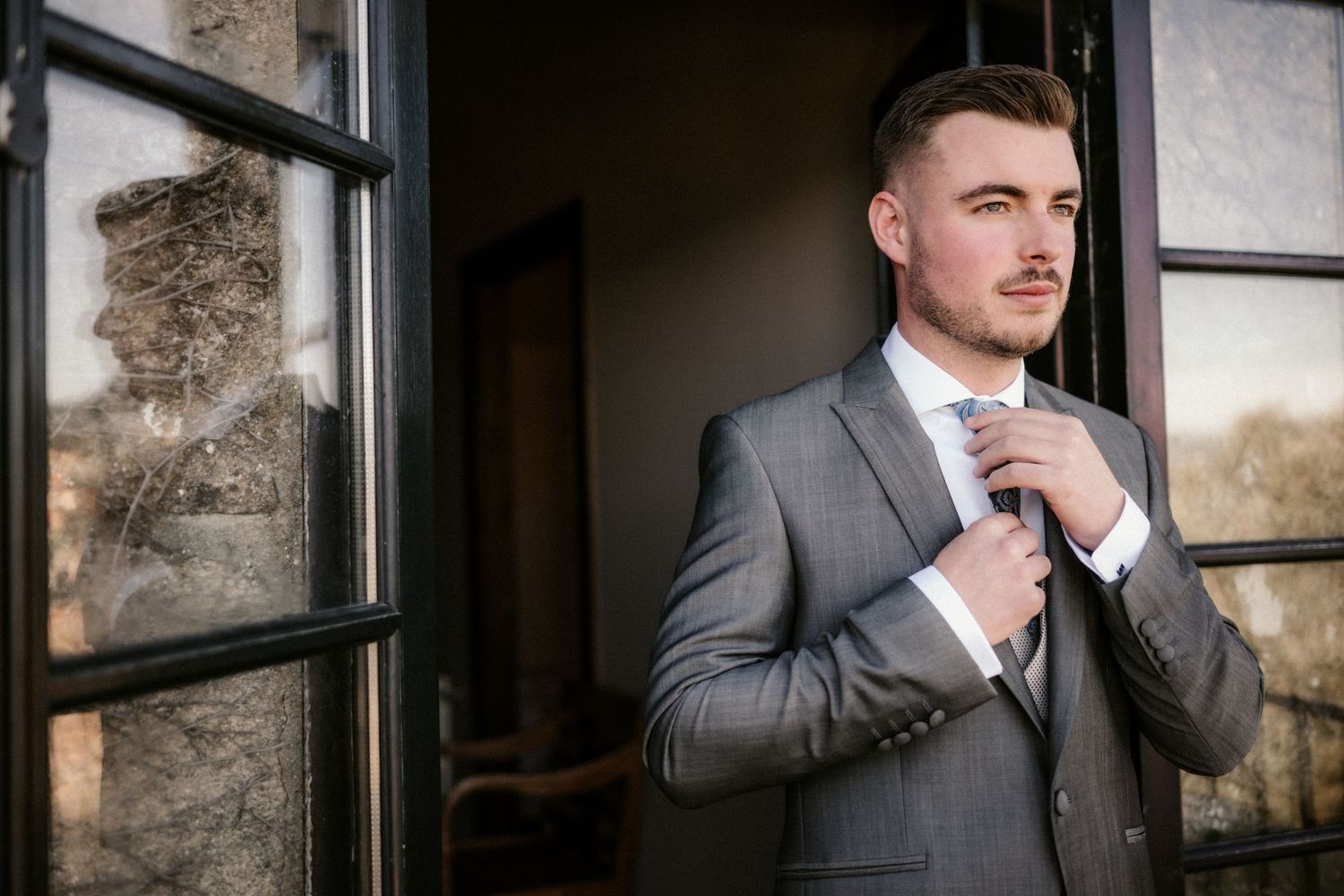 A man in a gray suit adjusts his tie while standing in front of a stone wall with window panes.