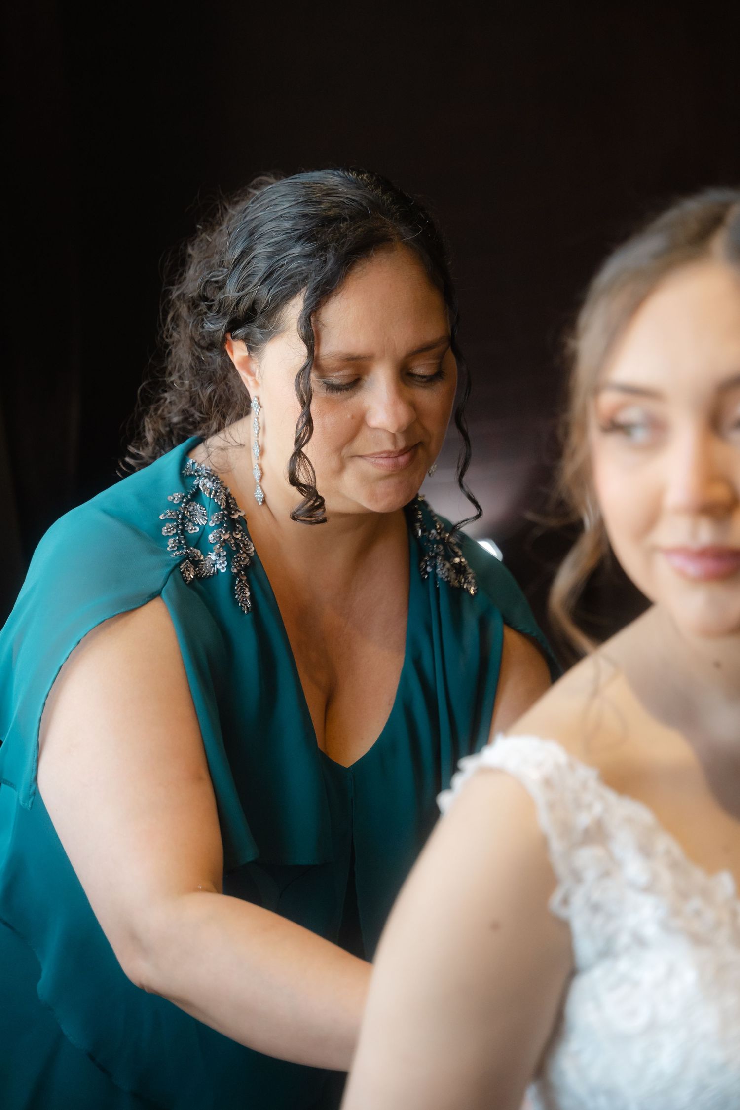 A person in a teal dress helps adjust a white lace wedding gown during preparation for a special occasion.