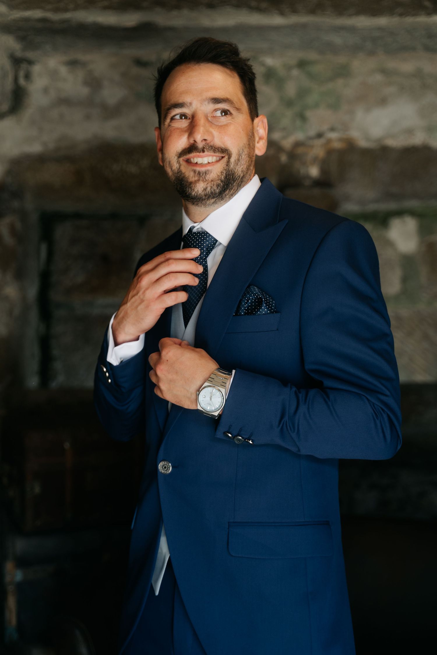 A person in a navy blue suit adjusts their tie and poses for formal portraits against a dark textured background.
