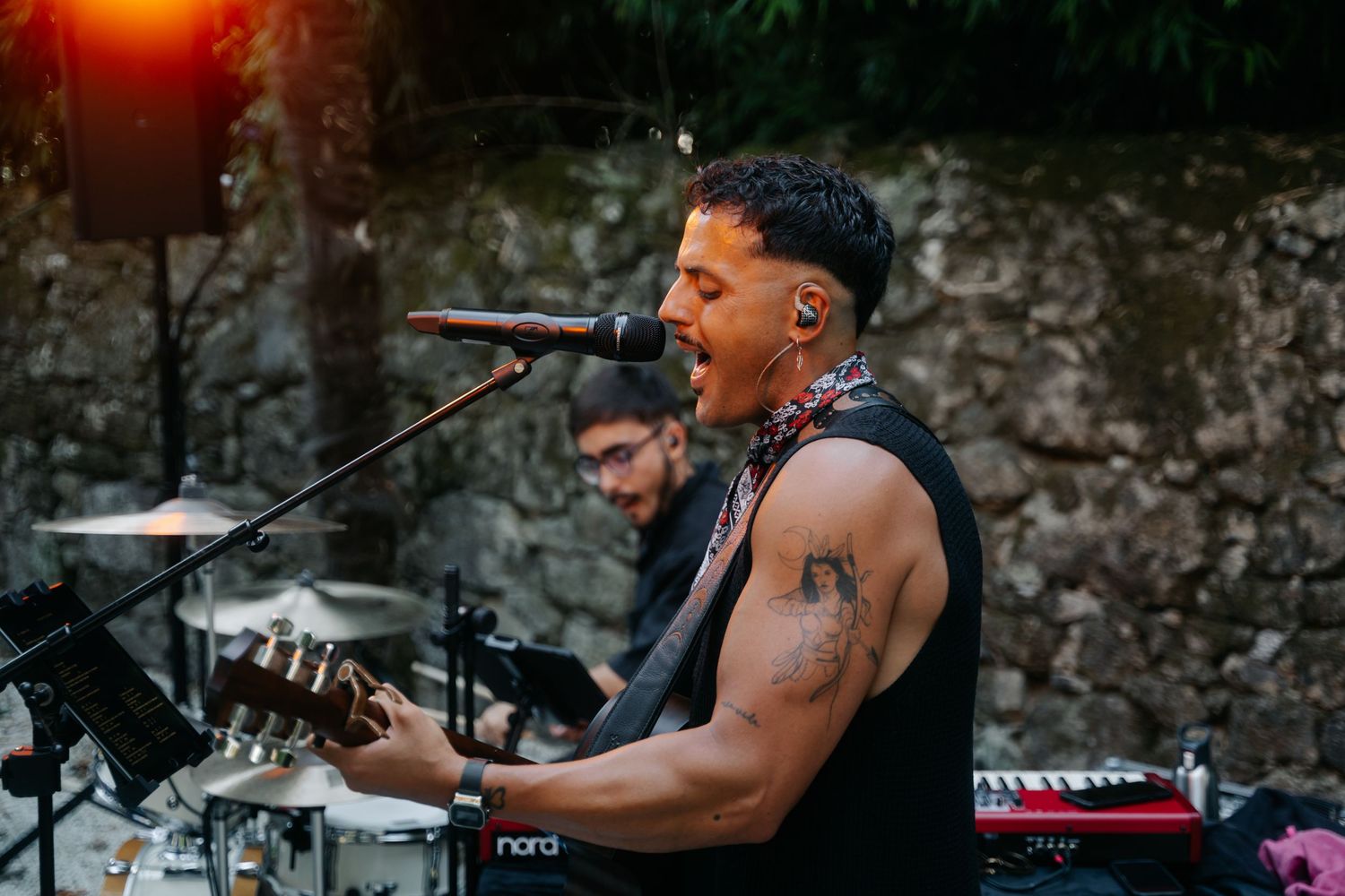 Band members play energetically on stage with microphones and instruments at a live outdoor performance.
