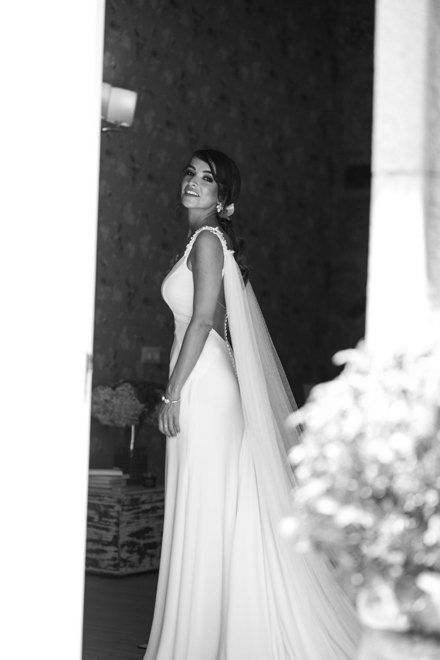 A bride in a flowing white wedding gown stands elegantly between classic white columns in a black and white photo.