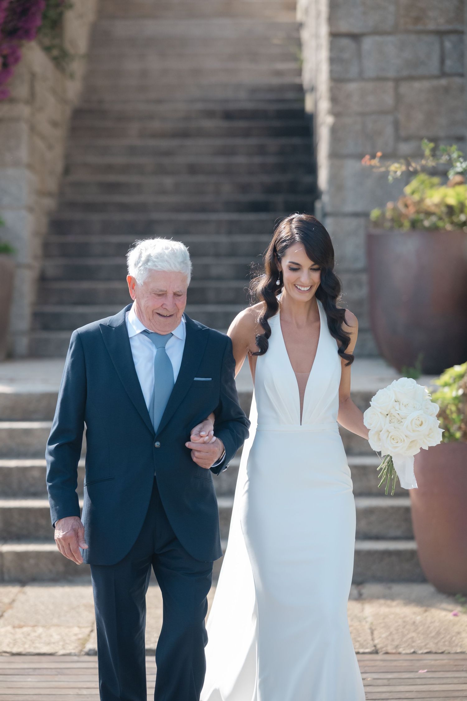 A bride in an elegant white gown descends stone stairs accompanied by an older gentleman in a navy suit.