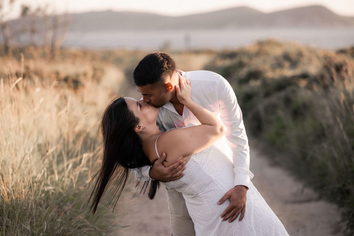 A couple in white wedding attire embraces lovingly in a golden field at sunset.