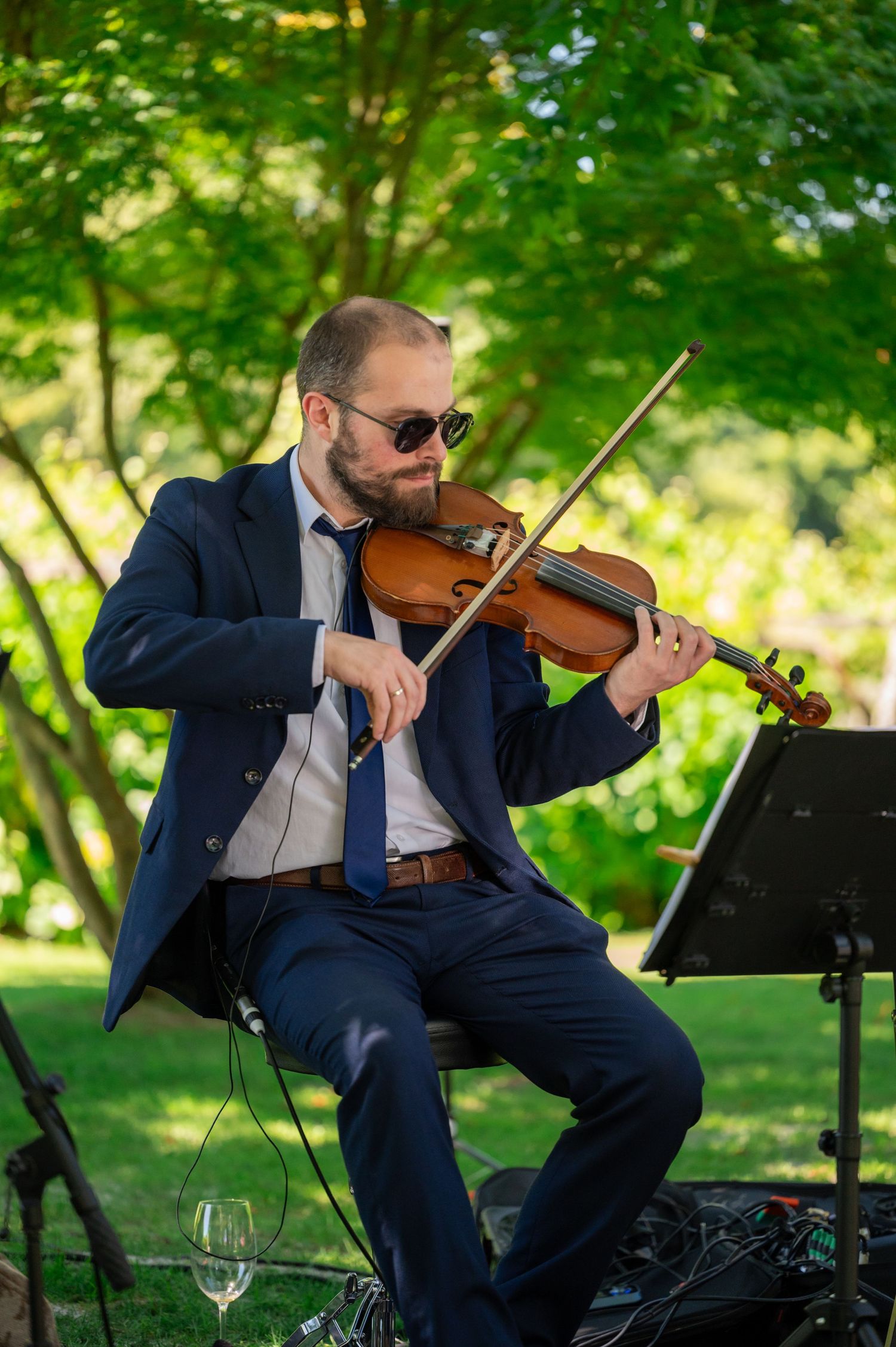 Violinist in a navy suit performs outdoors with a music stand against a verdant garden backdrop.