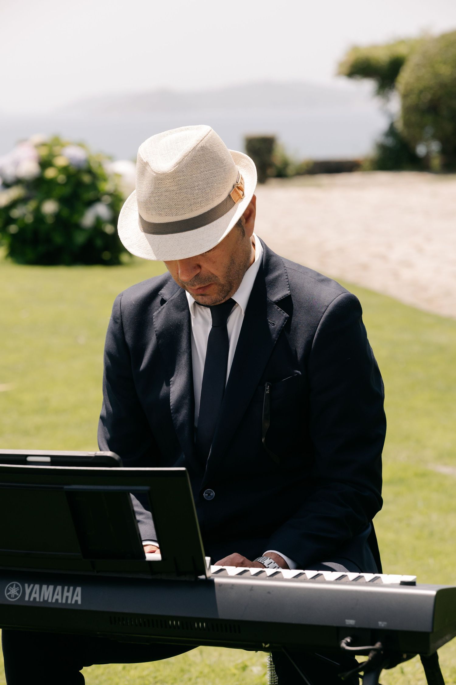 A pianist in a black suit and white hat plays a Yamaha keyboard outdoors on a sunny day.