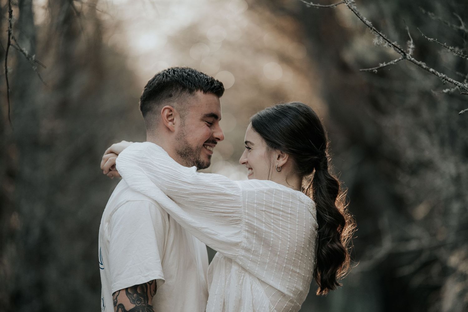 A romantic couple in white outfits embrace in a moody forest setting during a photo session.