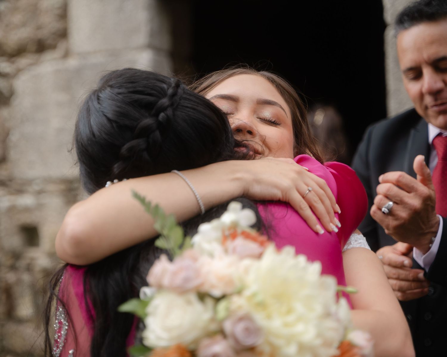 Wedding guests share emotional embraces during celebration by stone wall.