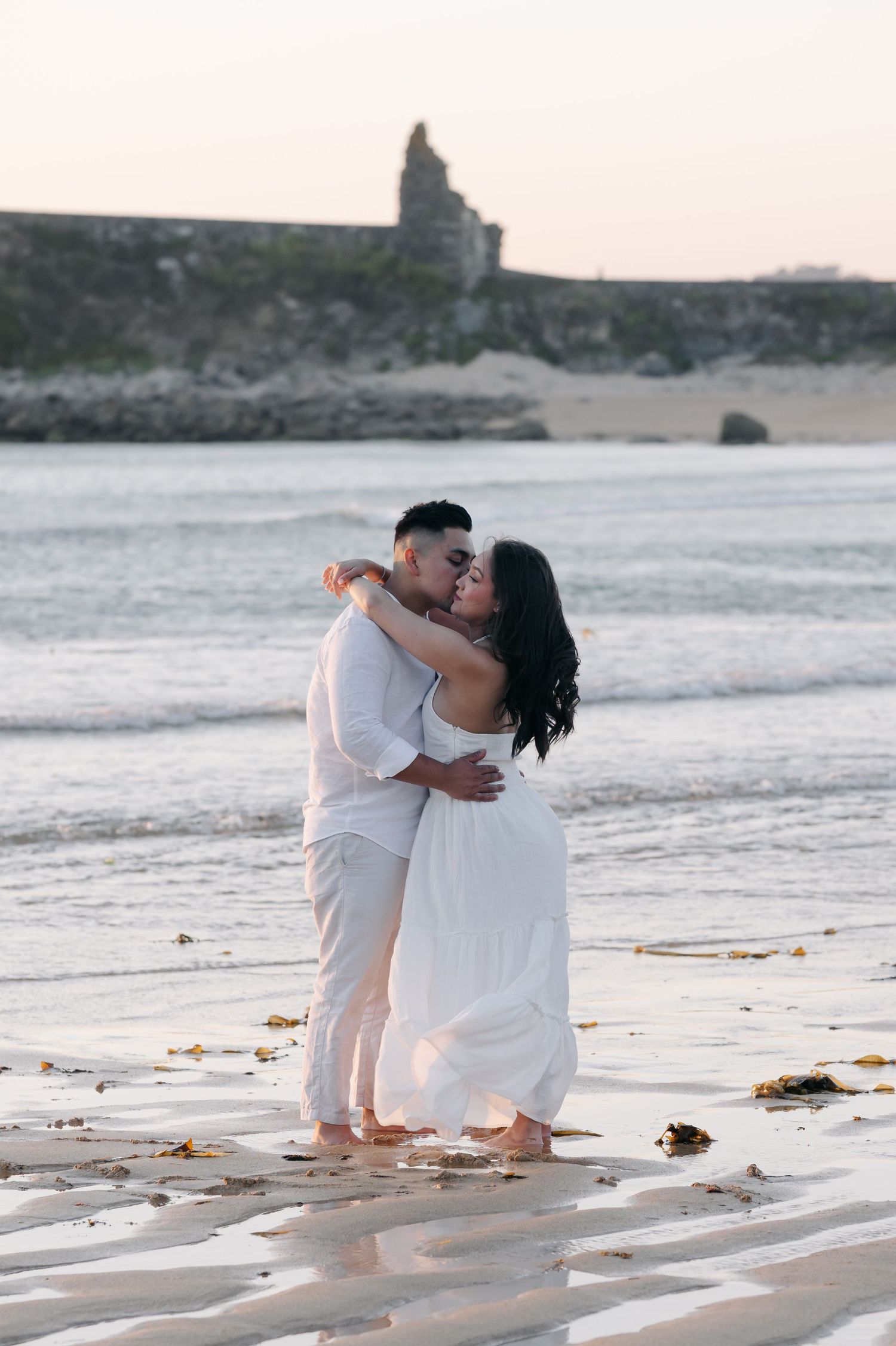 A couple embraces on a serene beach during sunset with waves gently lapping at the shore.