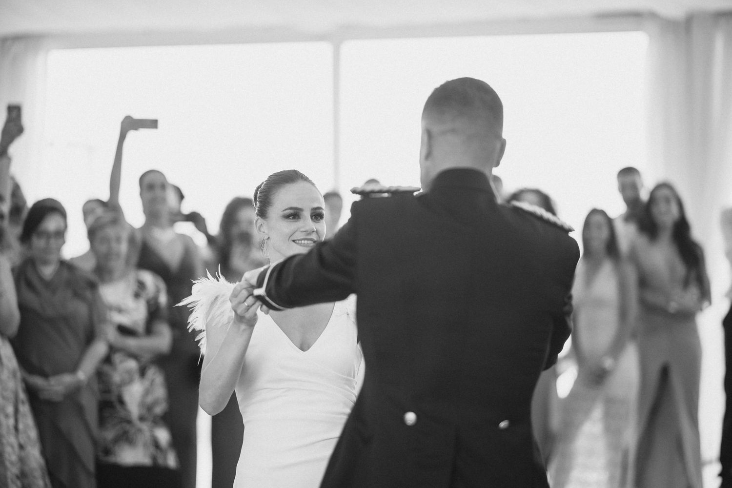 A couple shares their first dance at a wedding reception surrounded by guests in a bright indoor venue.