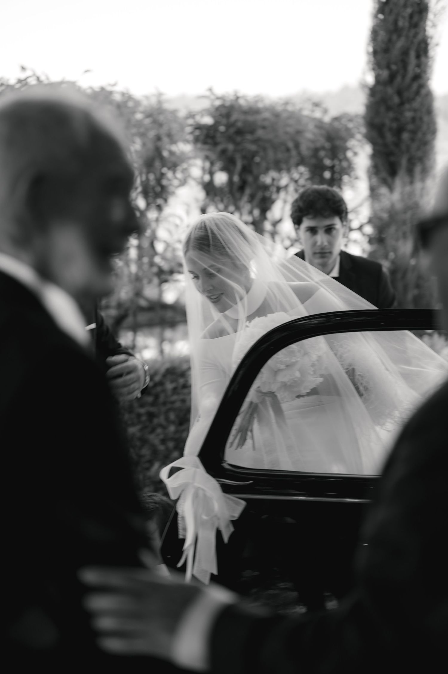 A black and white wedding photo shows a bride in a veil being helped into a car.