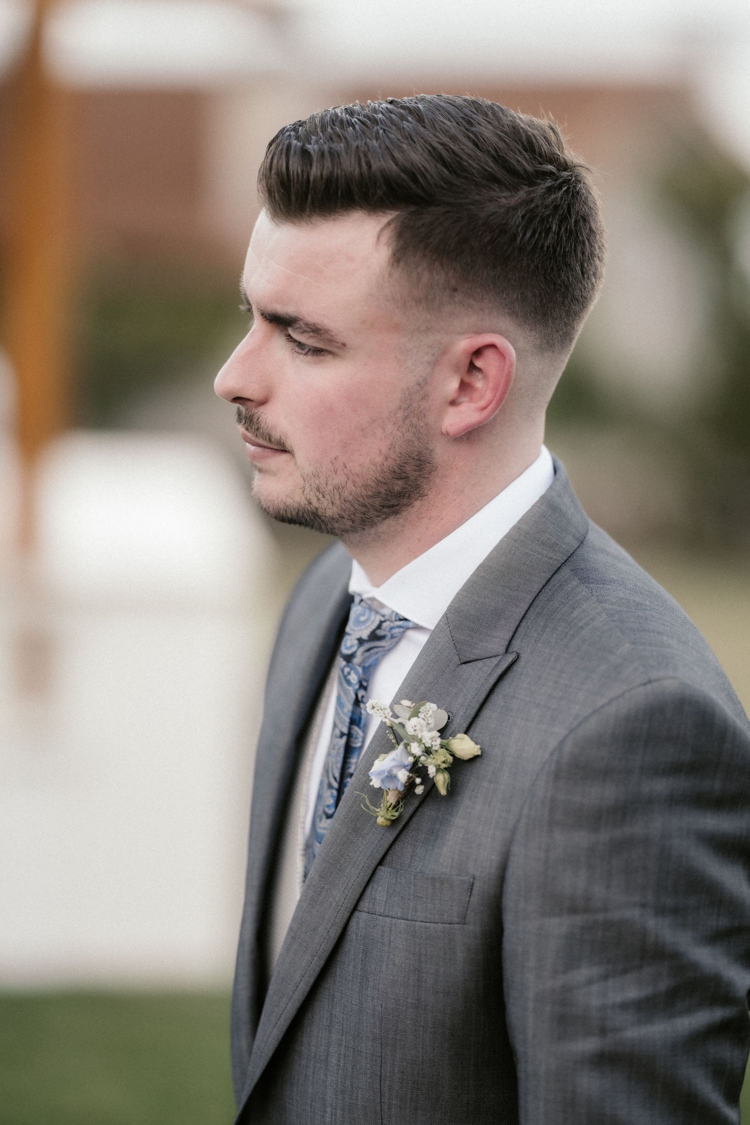 A groom in a grey suit and floral boutonniere stands in profile during an outdoor wedding.
