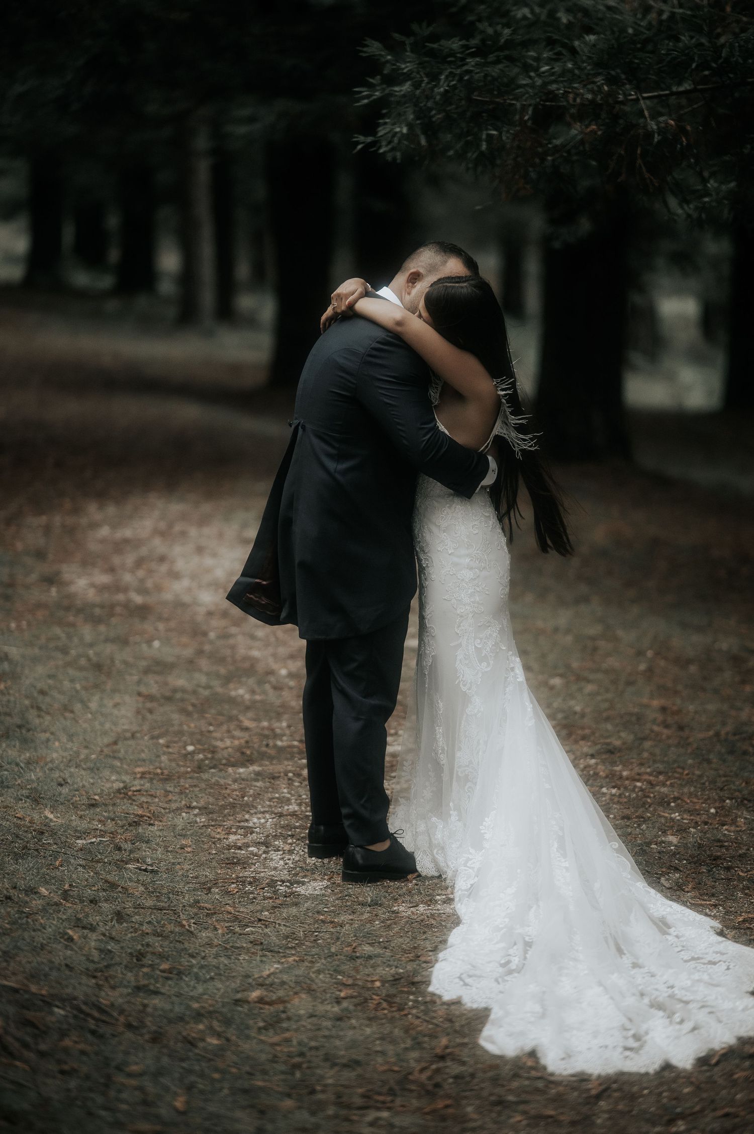 A romantic wedding couple embraces on a tree-lined path, with the bride's white lace dress trailing behind.