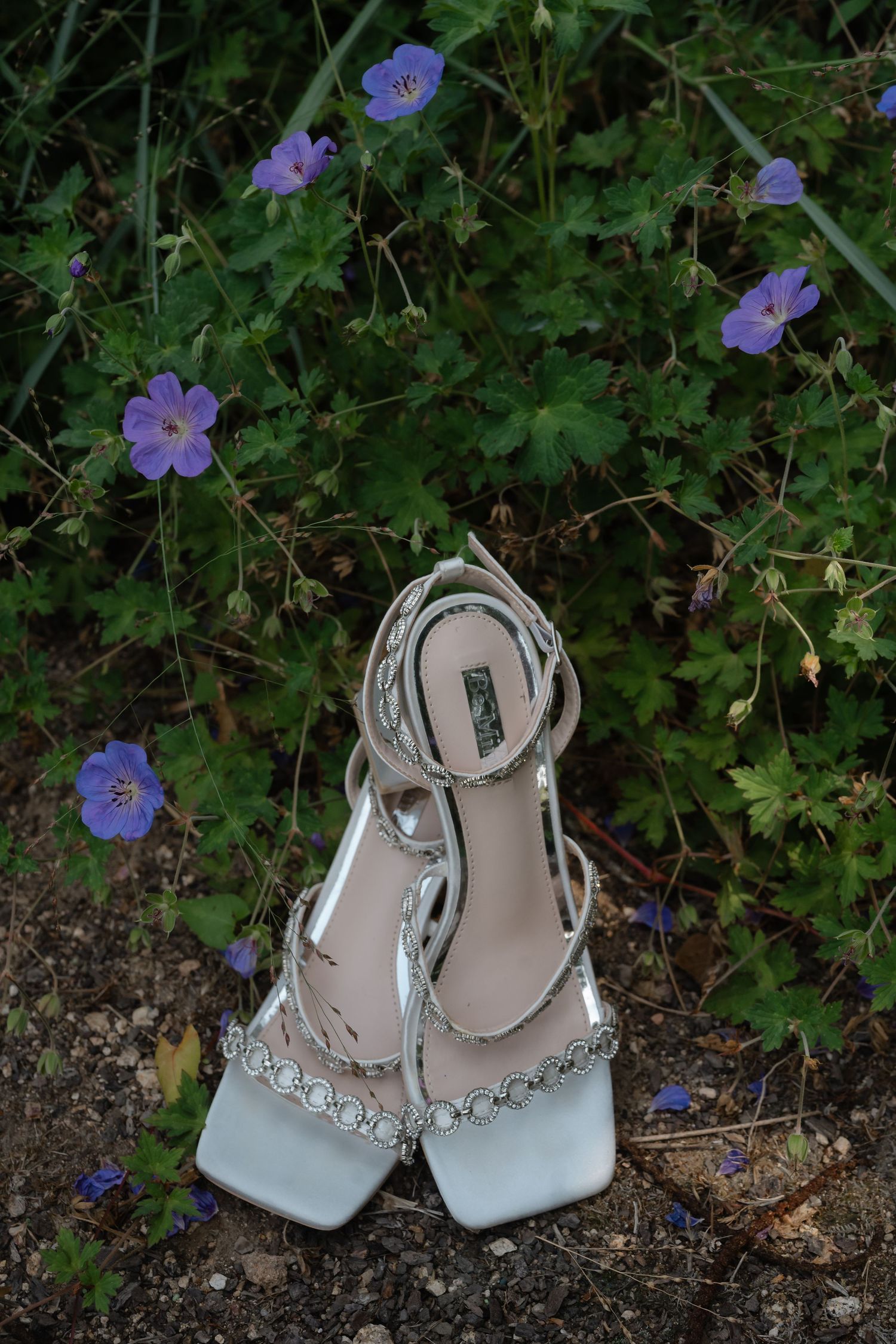 A pair of elegant white bridal shoes with crystal embellishments placed on the ground among purple wildflowers.