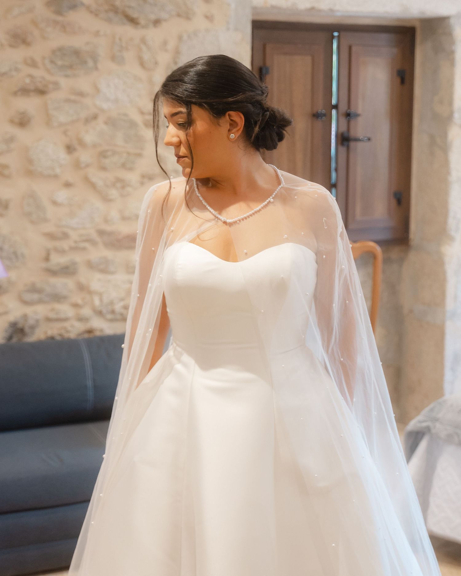 Flowing white wedding dress with delicate veil captured in soft natural light against stone wall.