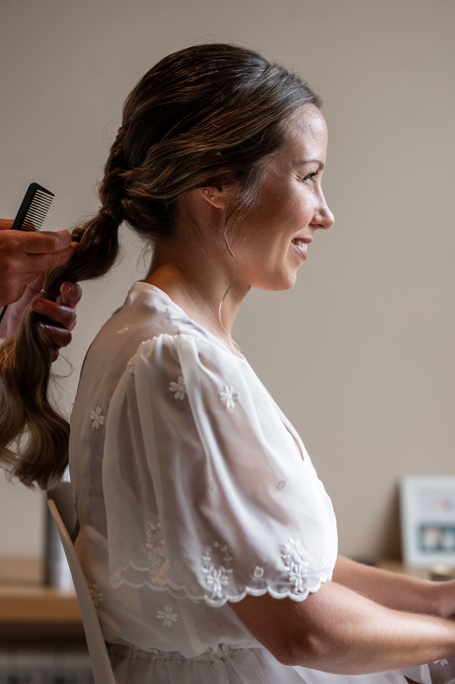 A person in a white lace dress smiles while having their long brown hair styled before a special occasion.