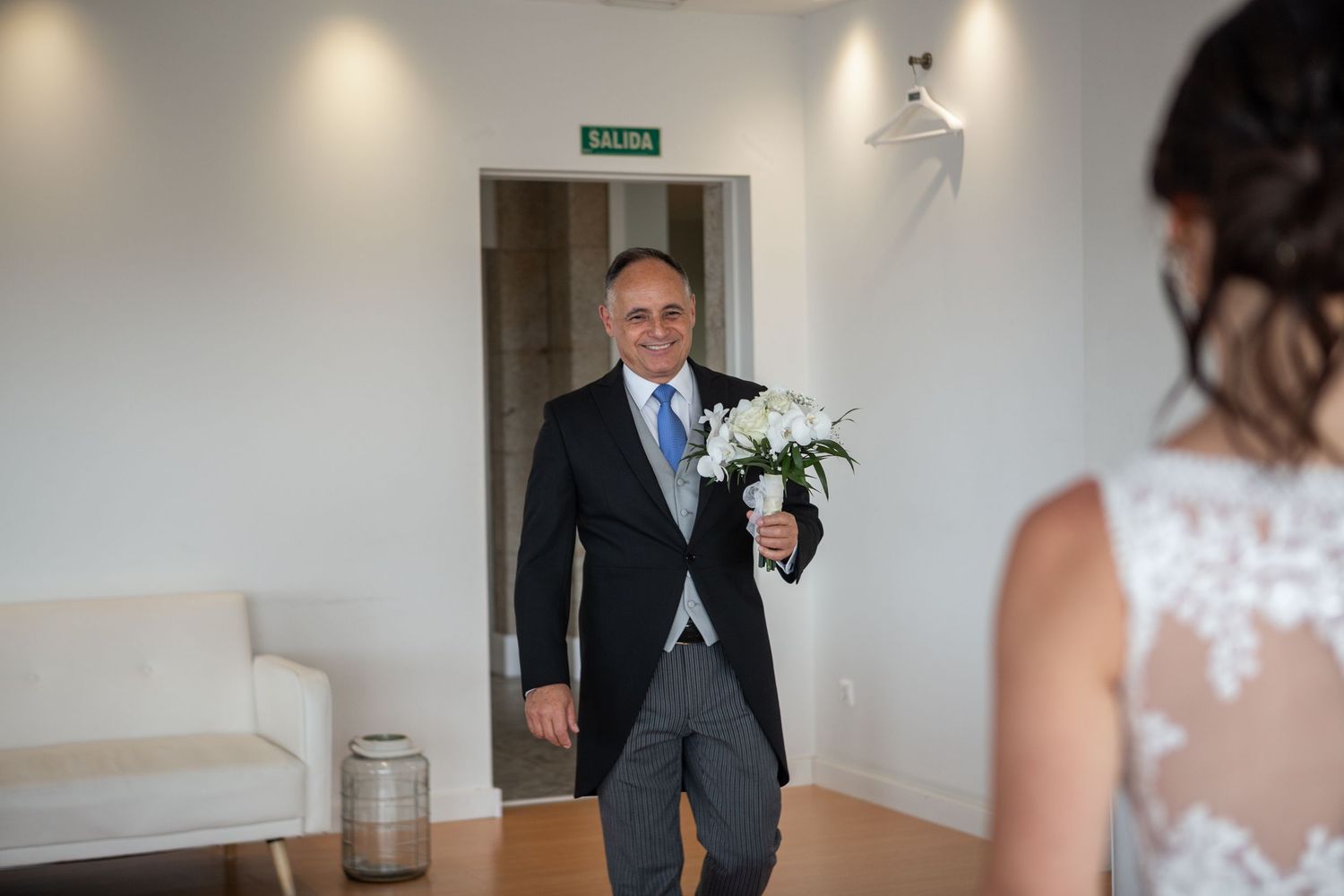 A person in a formal gray suit and tie holds a bouquet of white flowers in an indoor setting.