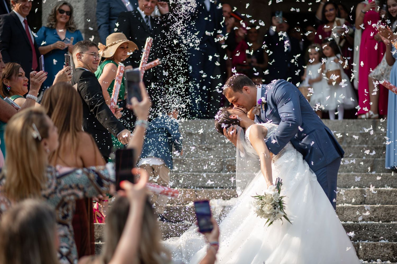 Wedding party exits church while guests throw confetti in celebration on the stone steps.