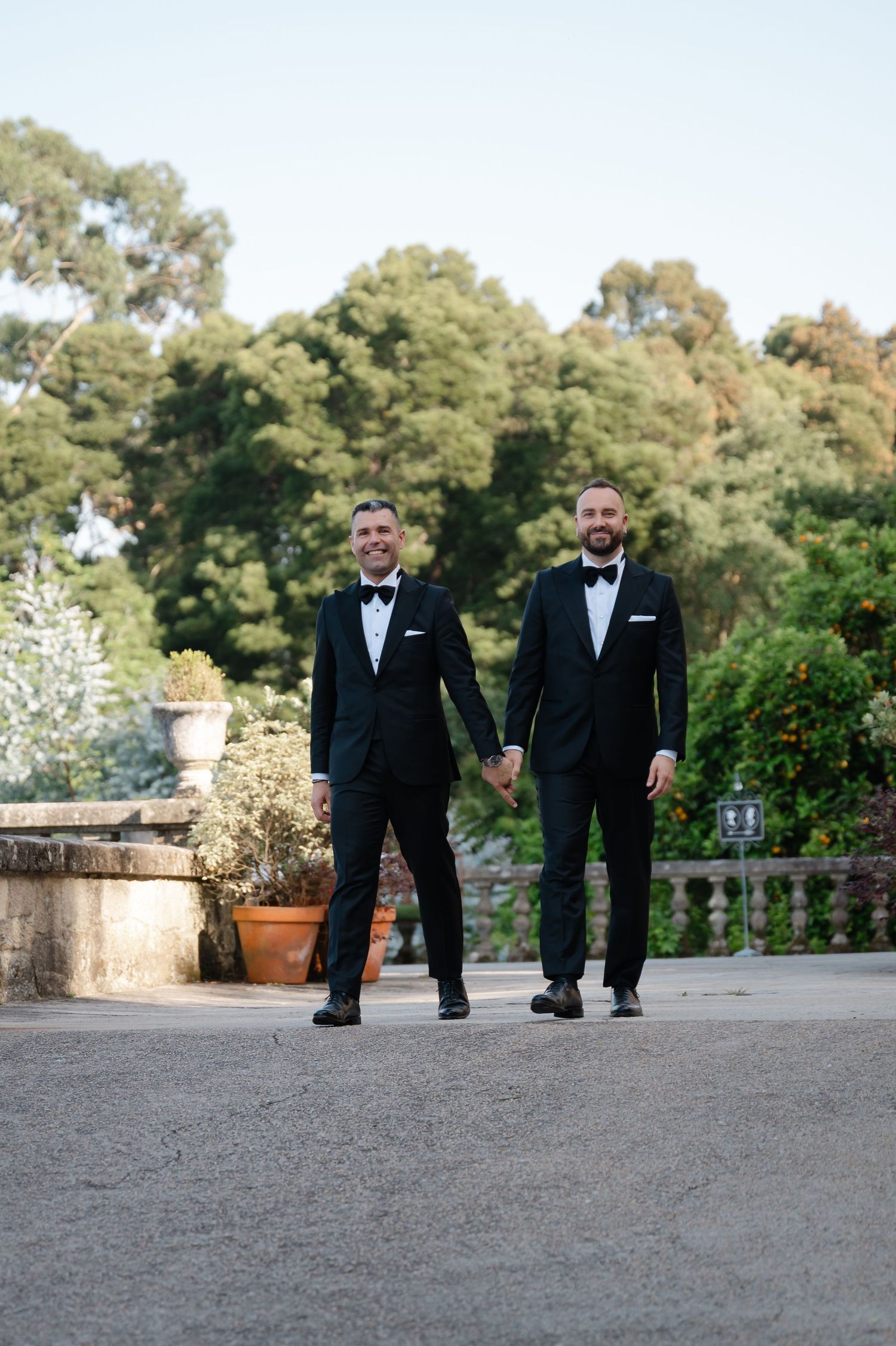 Two people in black tuxedos walking together on a stone patio with trees in the background.