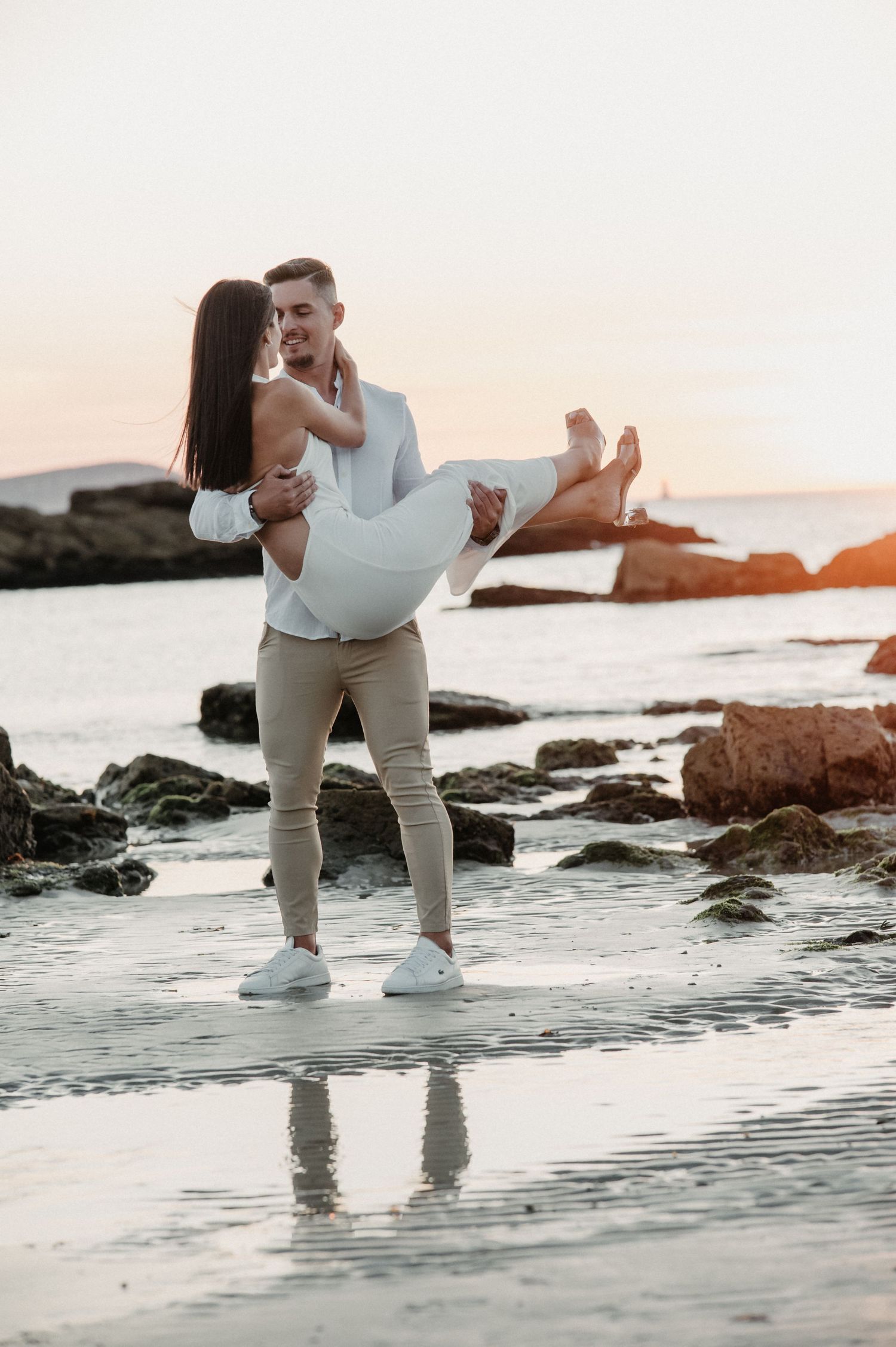 A romantic couple embraces on a rocky beach at sunset with waves lapping at their feet.