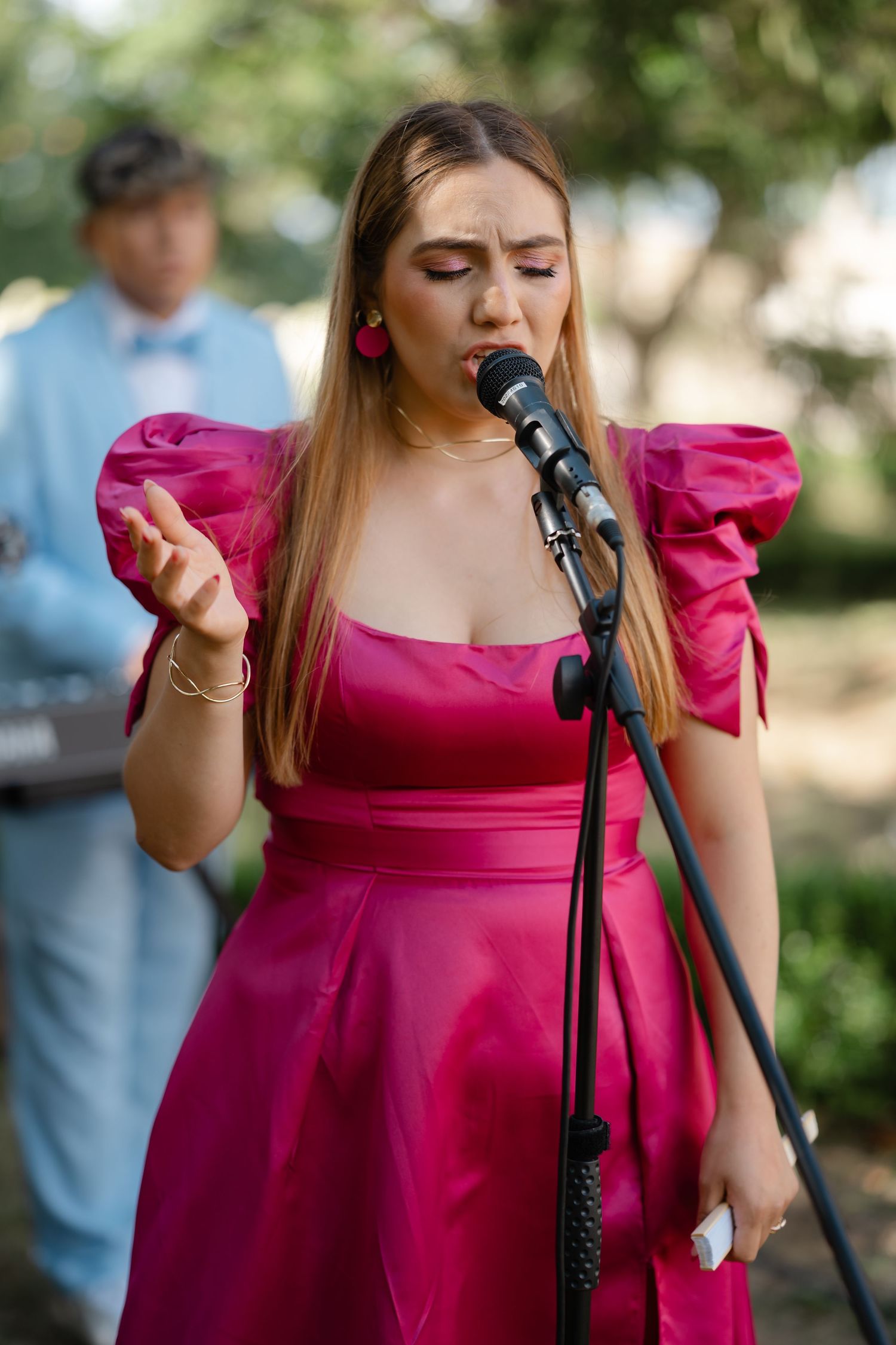 A performer in a vibrant pink dress sings passionately into a microphone during an outdoor performance.