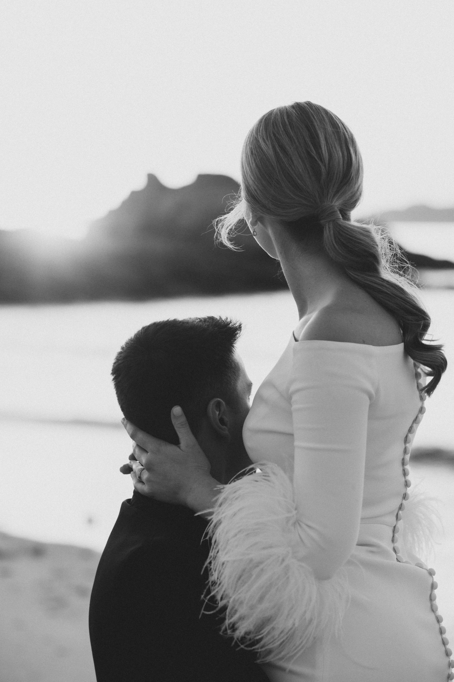 Black and white portrait of newlyweds embracing against coastal landscape.