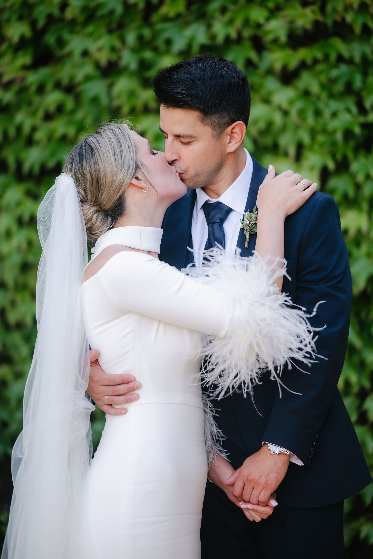 Bride in white feathered dress and groom in navy suit share tender moments in garden wedding photoshoot.