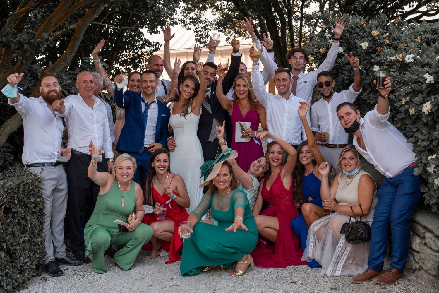 Large wedding party group photo with guests wearing colorful attire under trees outdoors.