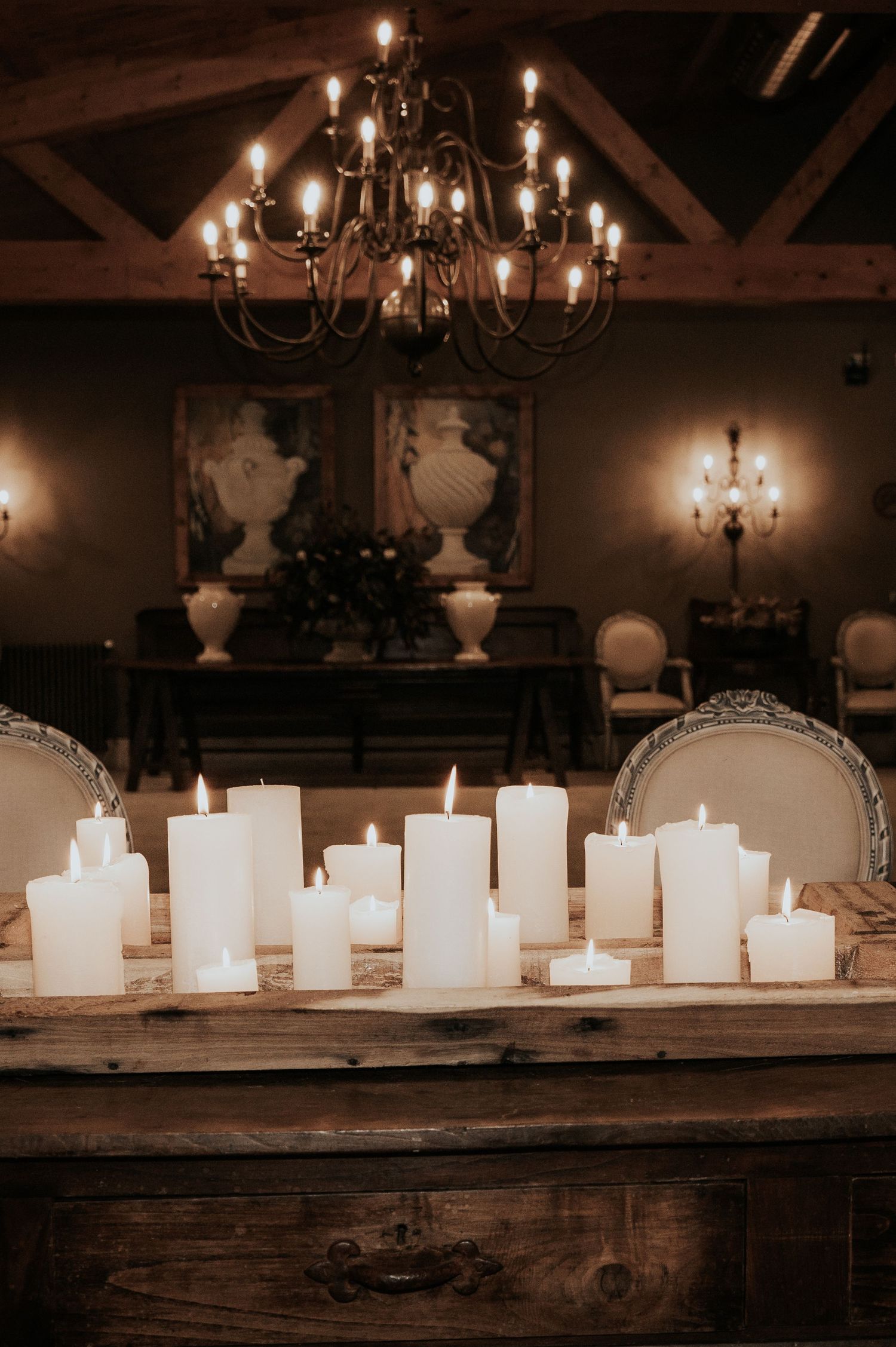 White pillar candles arranged on rustic wooden table with elegant chandelier and ornate decor in dimly lit room.
