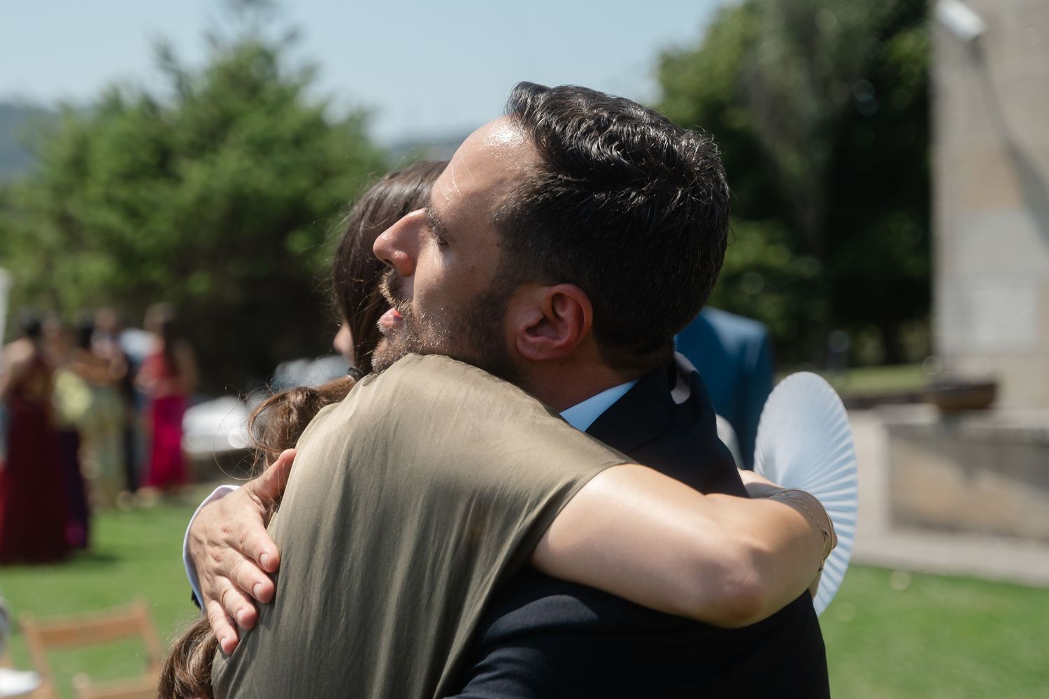 Two people share an emotional embrace outdoors in a garden setting during a special occasion.