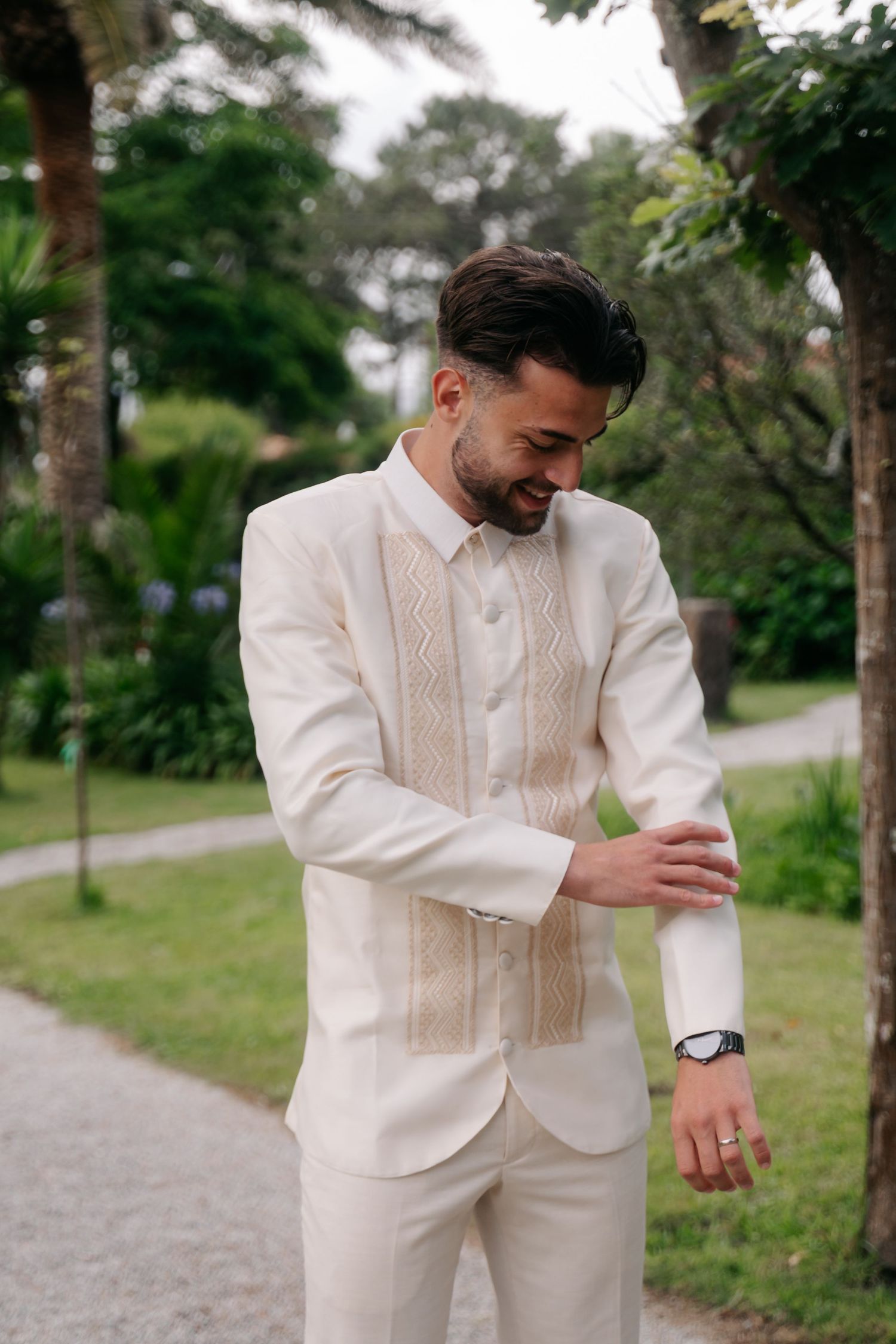 Groom in an elegant cream-colored suit with textured vest checks his watch while standing on garden path.