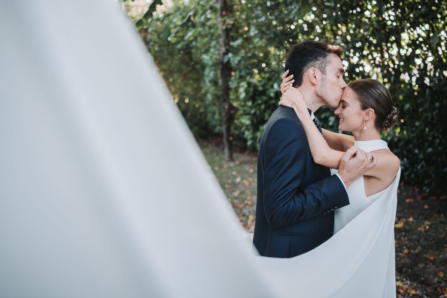 A romantic wedding couple shares a tender kiss outdoors while her white veil flows in the breeze.