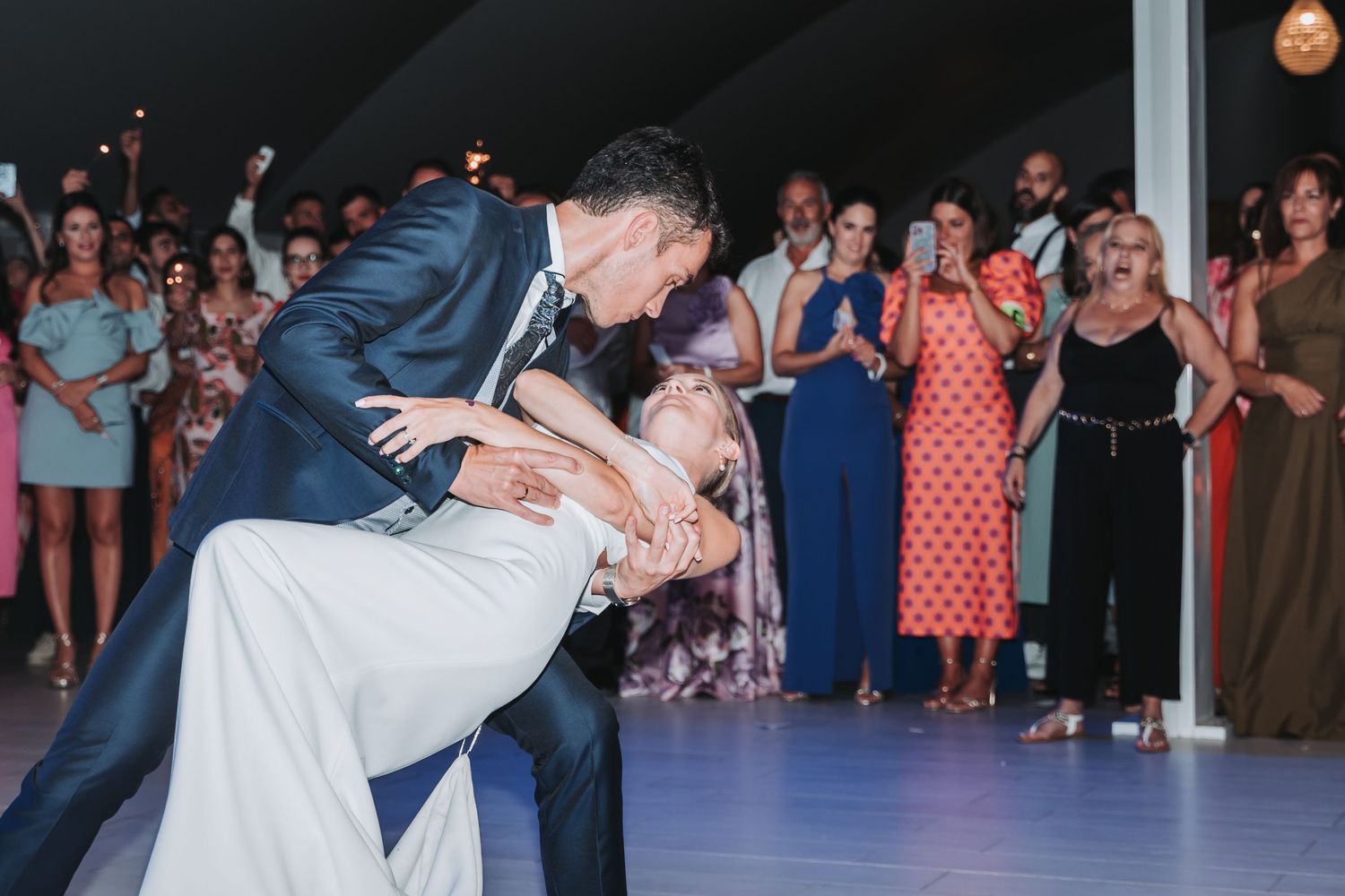 A couple performs a dramatic dip during a wedding dance on a lit dance floor surrounded by guests.