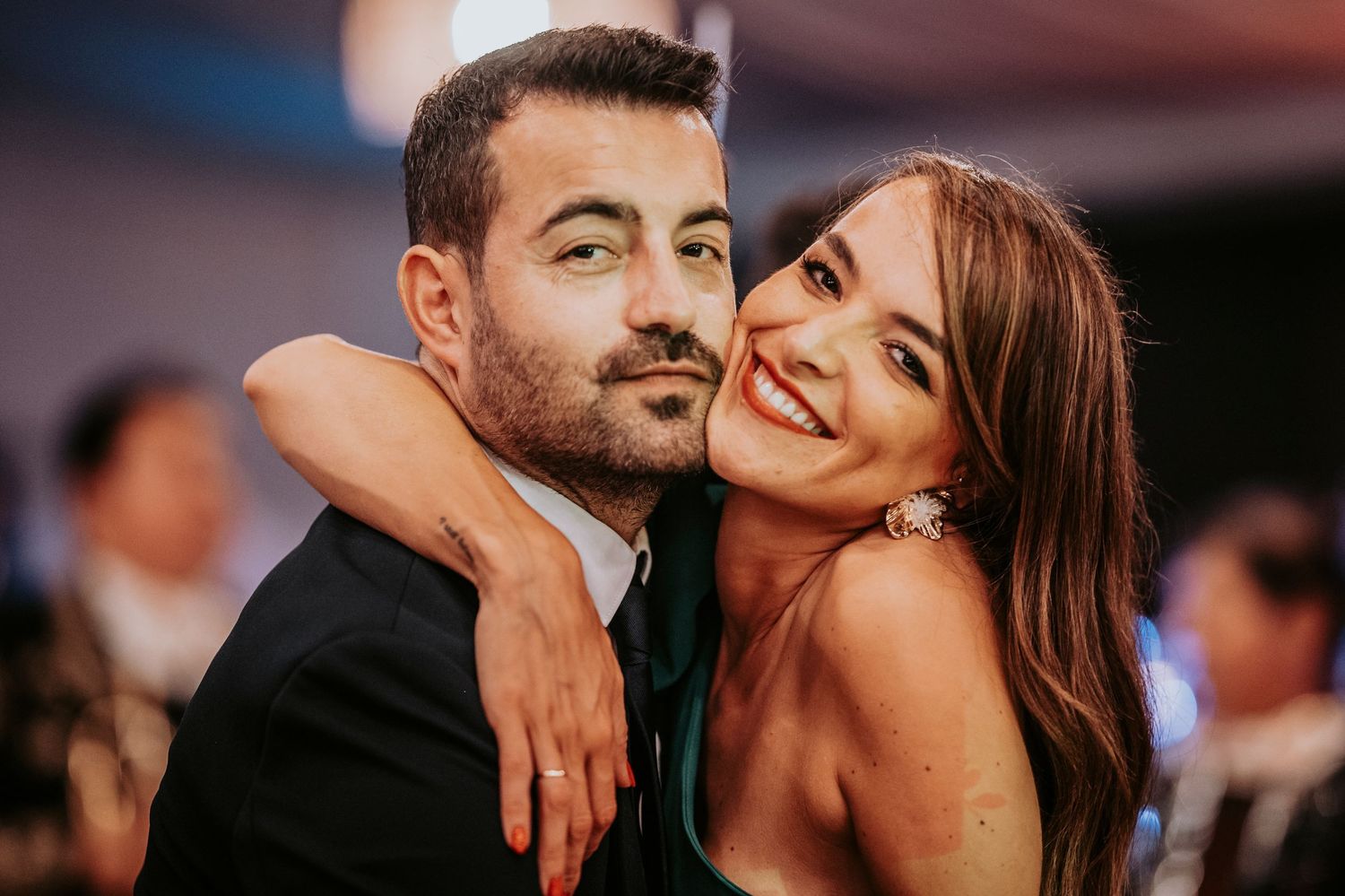 A happy couple embraces at an evening event, with the woman draping her arm around her partner's shoulder.