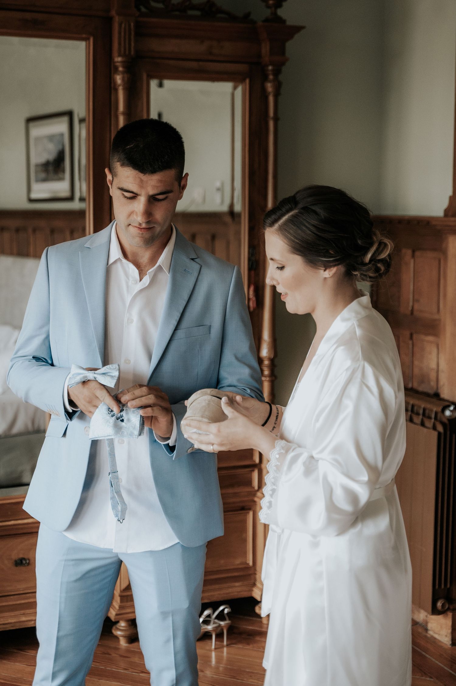 A couple getting ready before their wedding, with one wearing a light blue suit and the other in a white robe.
