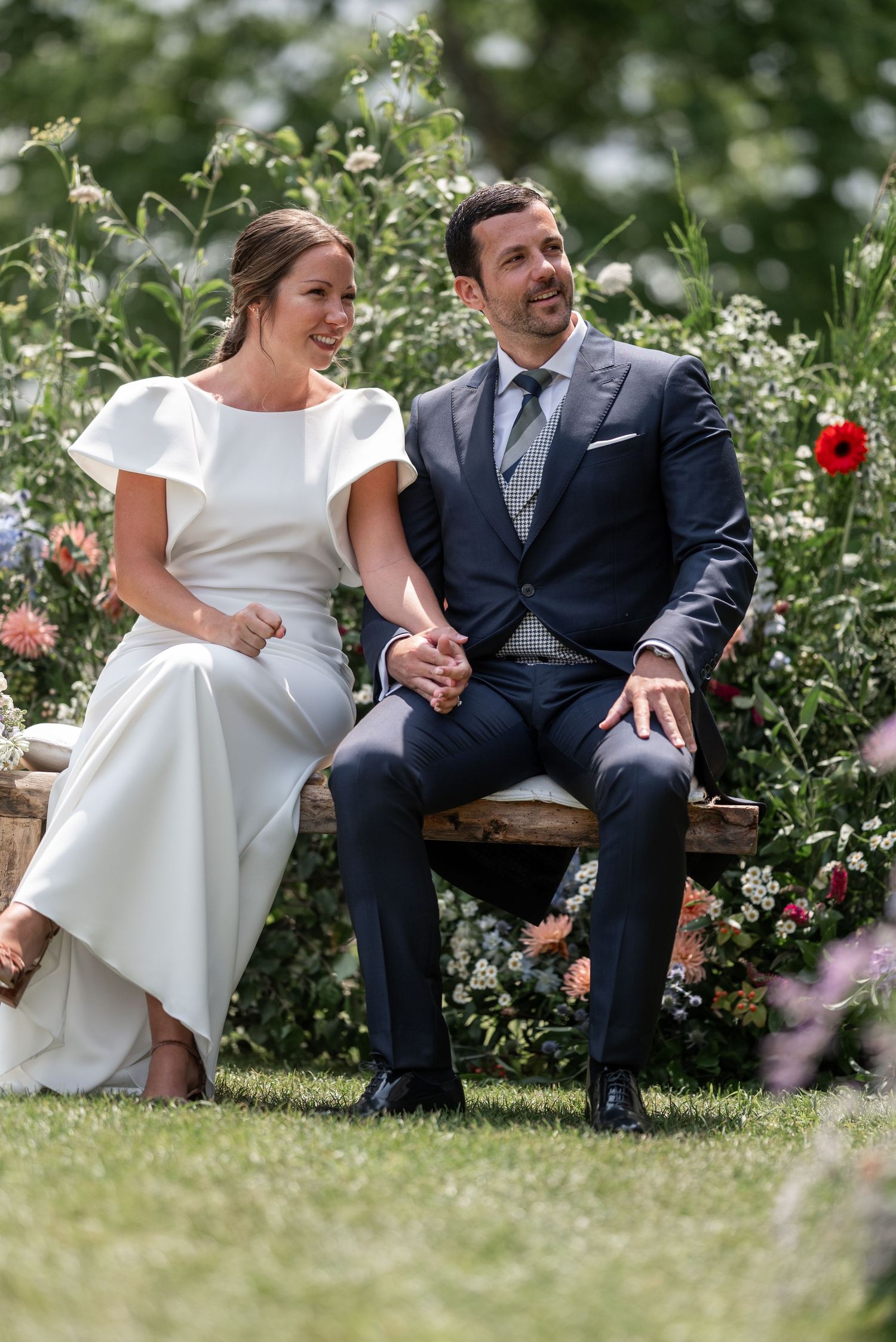 A happy couple shares a joyful moment together on a wooden bench surrounded by blooming flowers in a garden setting.
