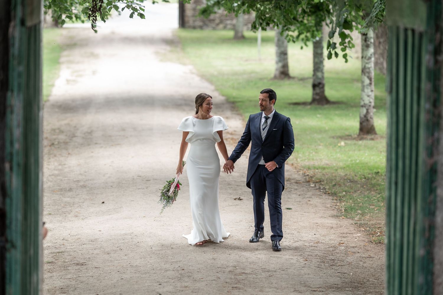 A couple walks together on a tree-lined path in a park setting during what appears to be a wedding celebration.