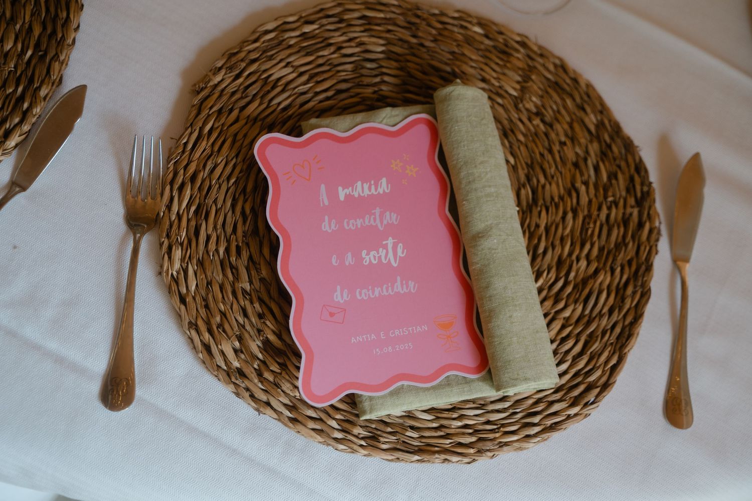 Woven rattan charger plates with wooden utensils and pink decorative cards arranged on white surface.