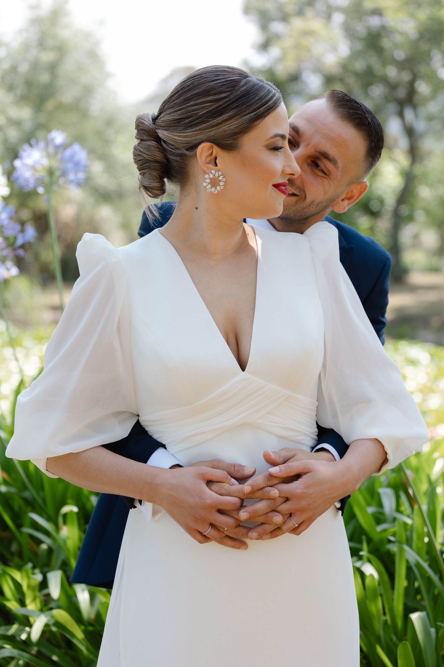 Bride in elegant white dress and groom in navy suit embrace tenderly in an outdoor garden setting.