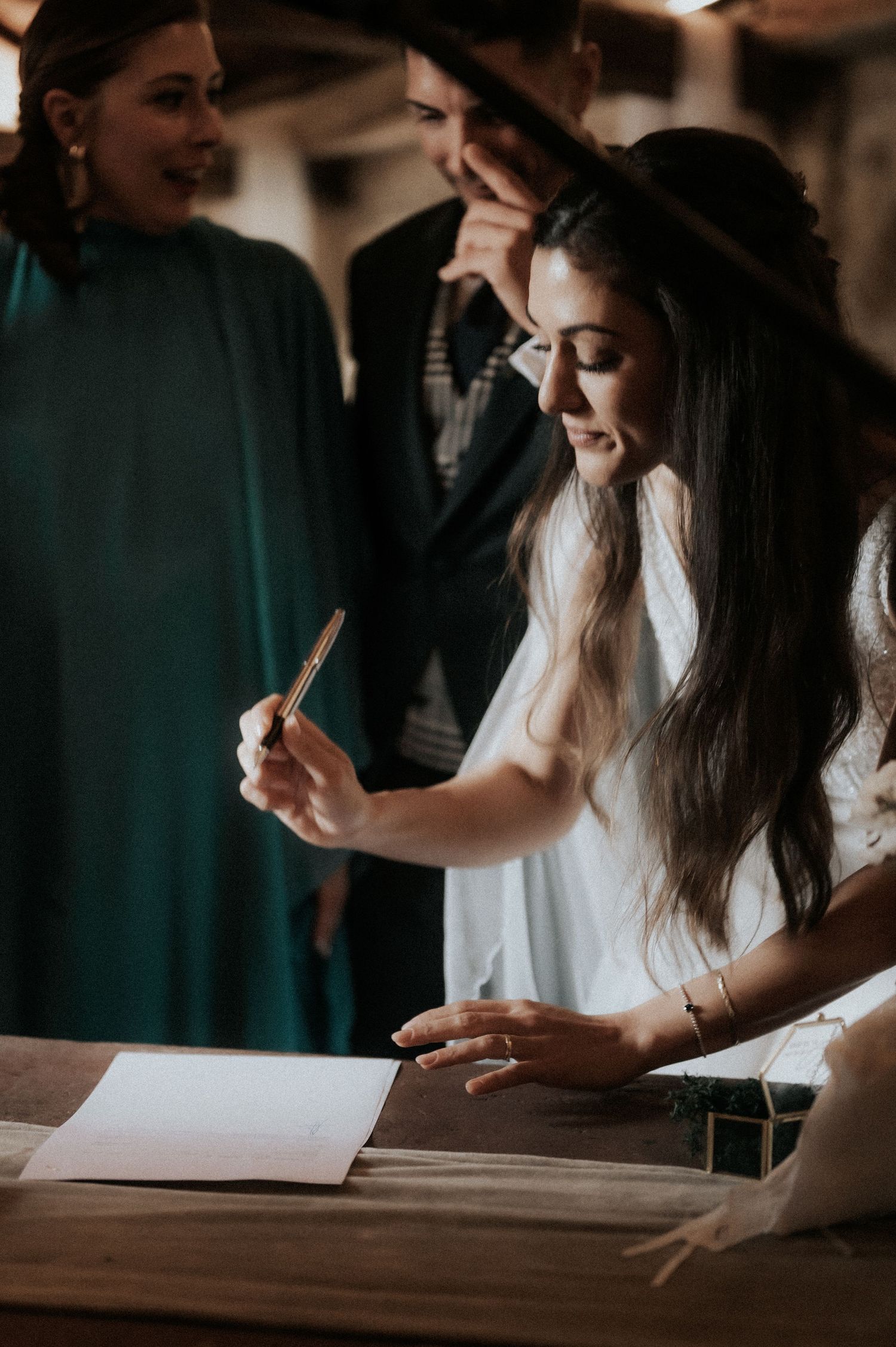 A person in a white dress signs a document at a wooden table while others look on in warm lighting.