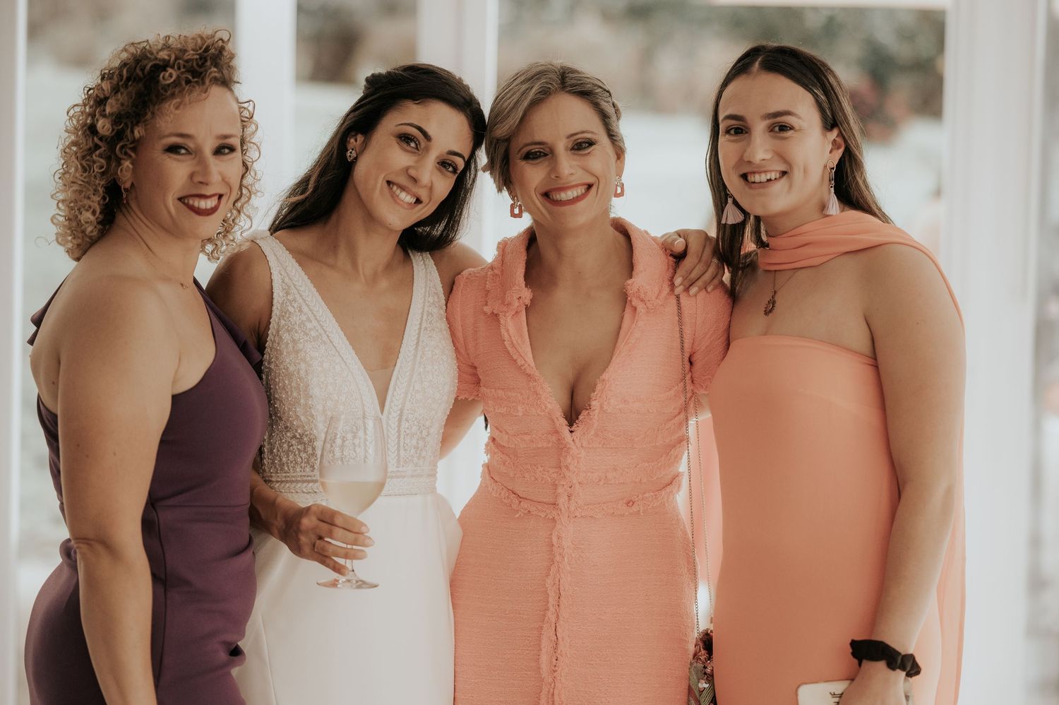 A group of bridesmaids in peach and purple dresses smile together in a bright indoor setting.