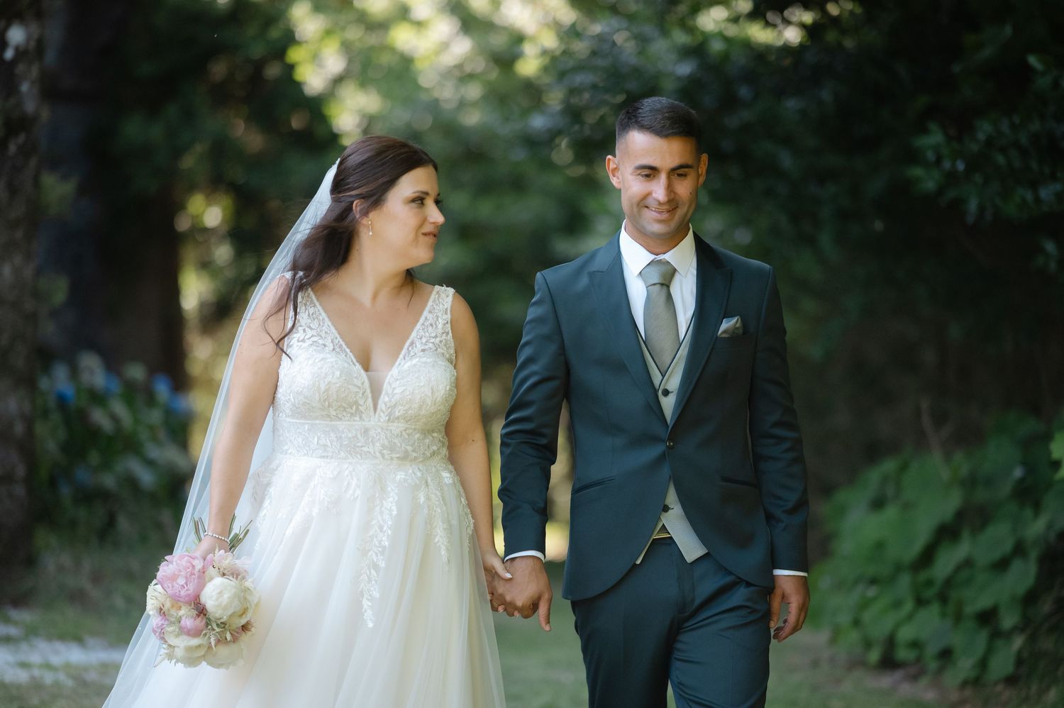 A couple in wedding attire walks together through a lush garden setting during their outdoor wedding photoshoot.