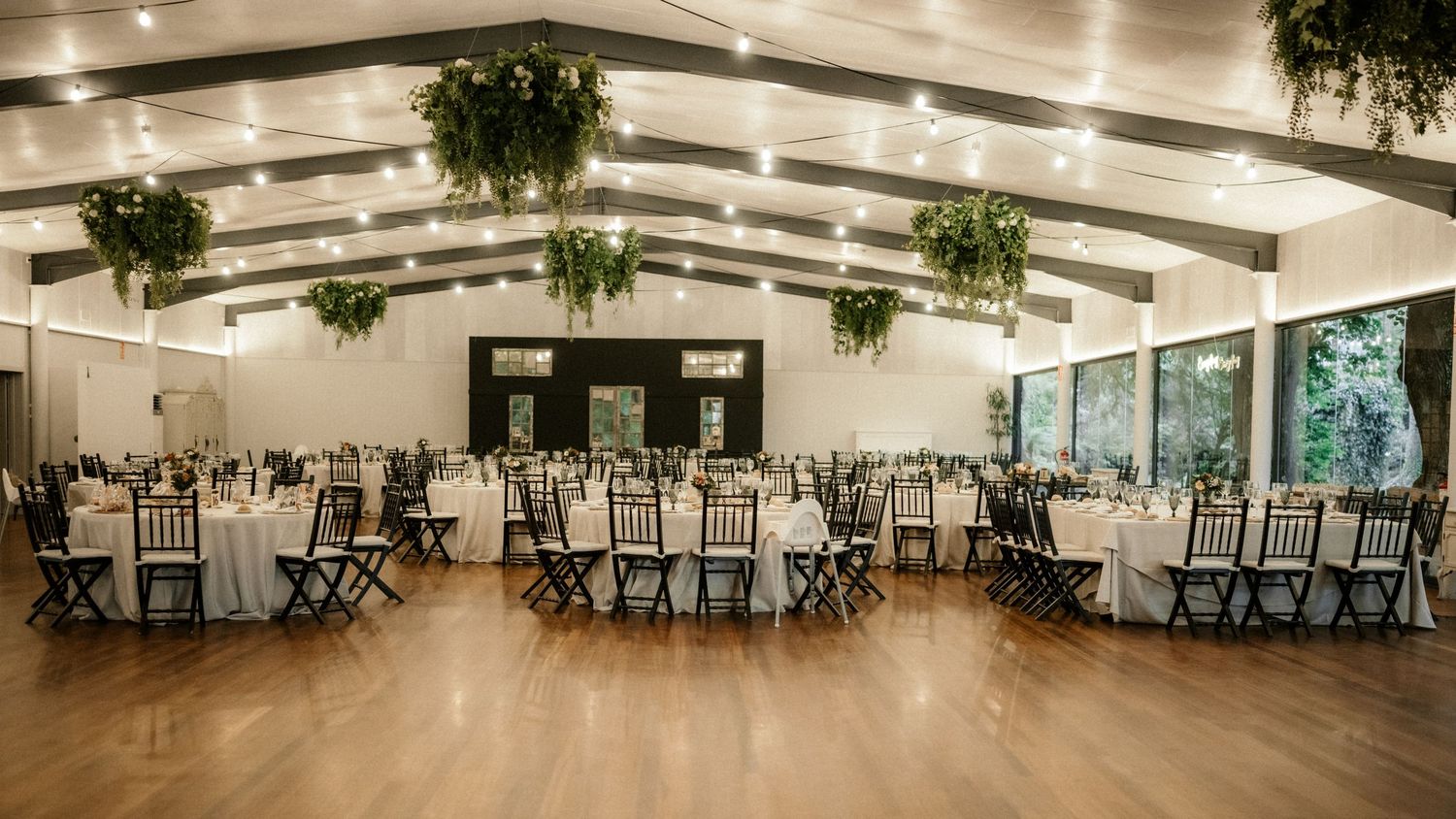 A panoramic view of an elegant banquet hall with round tables, white chairs, and hanging greenery decorations.