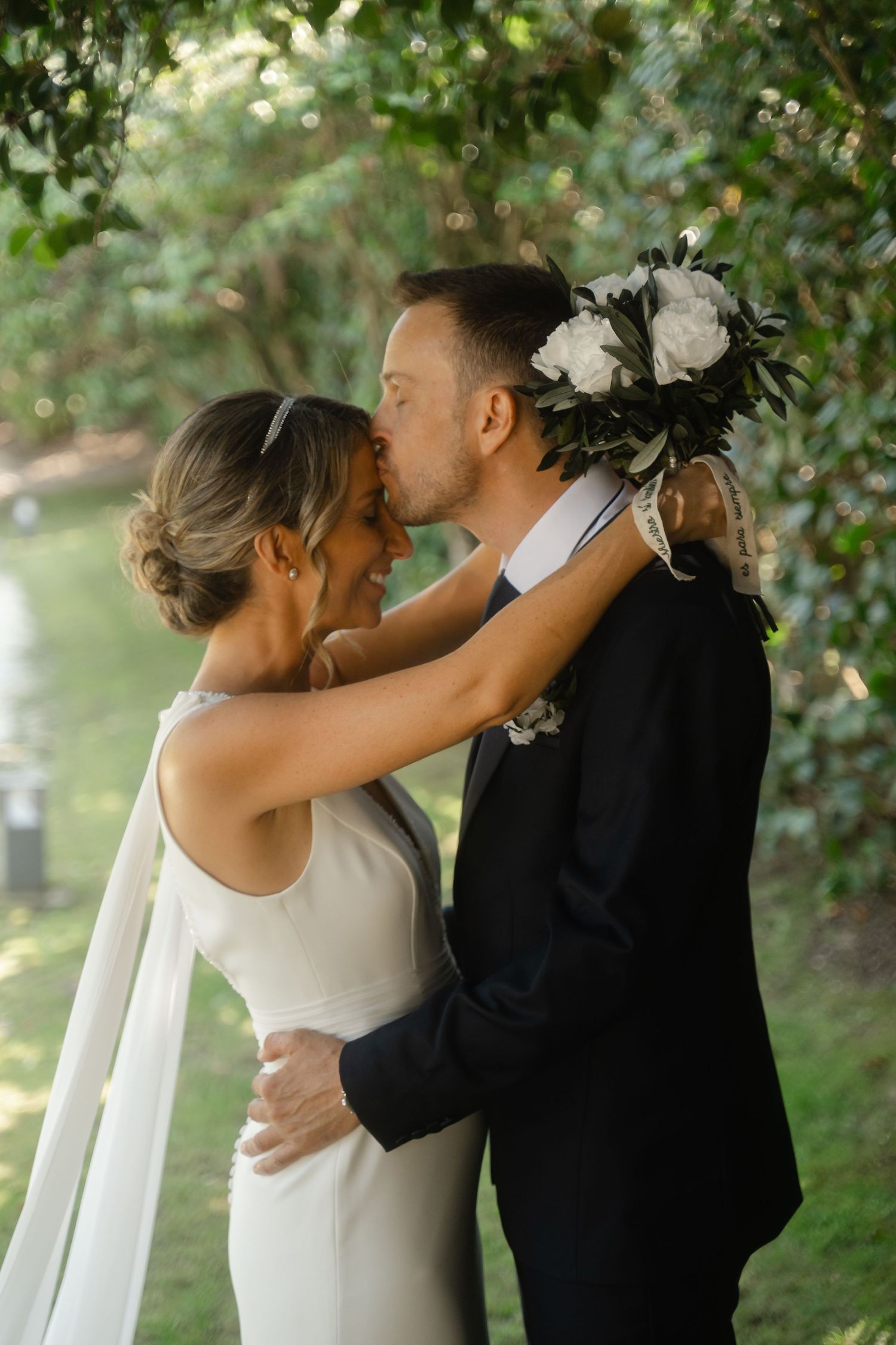 A romantic wedding moment captured as a couple embraces under green foliage wearing elegant white and black attire.