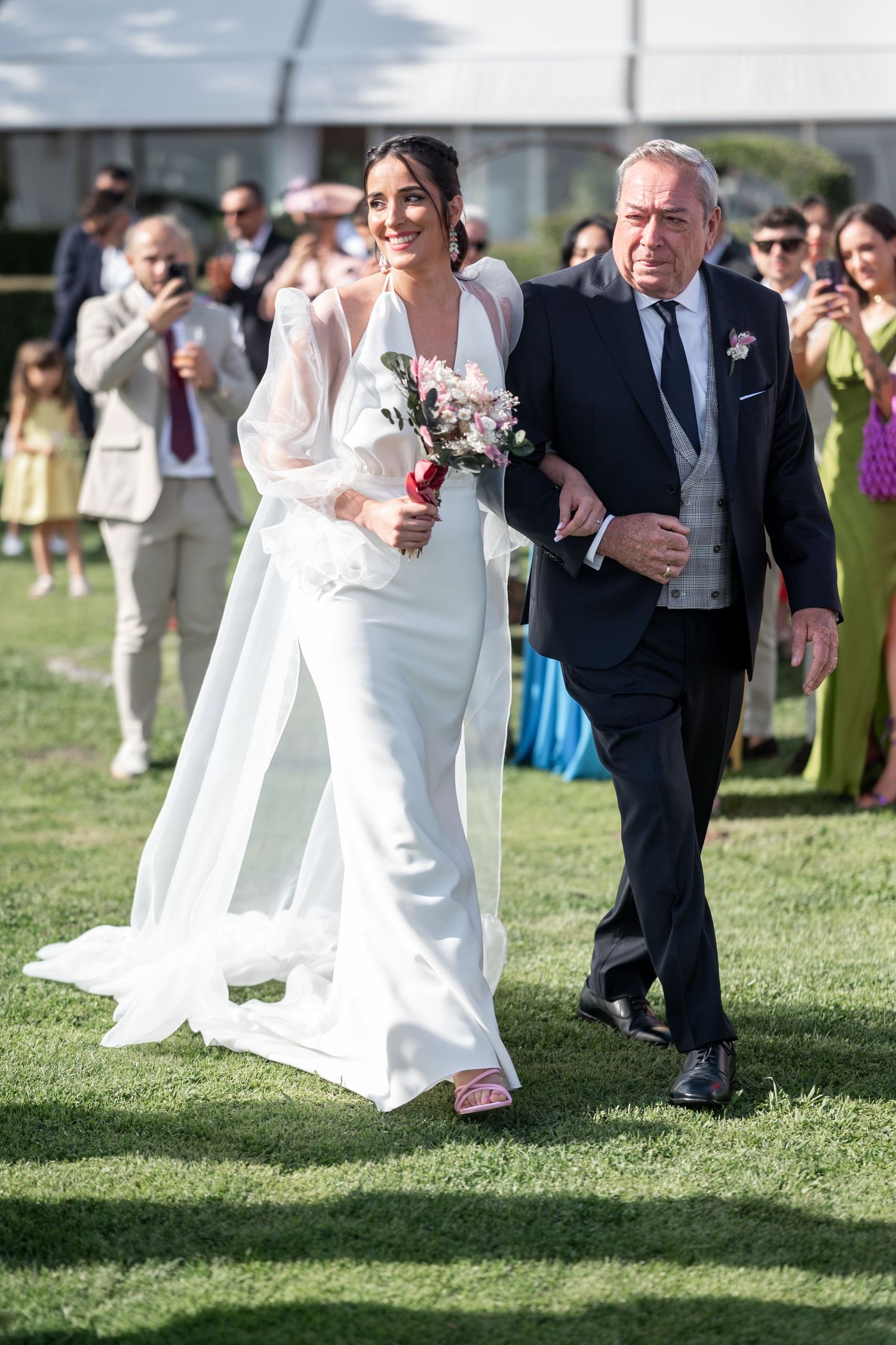 A bride in an elegant white dress walks down an outdoor aisle accompanied by an older gentleman in a formal suit.