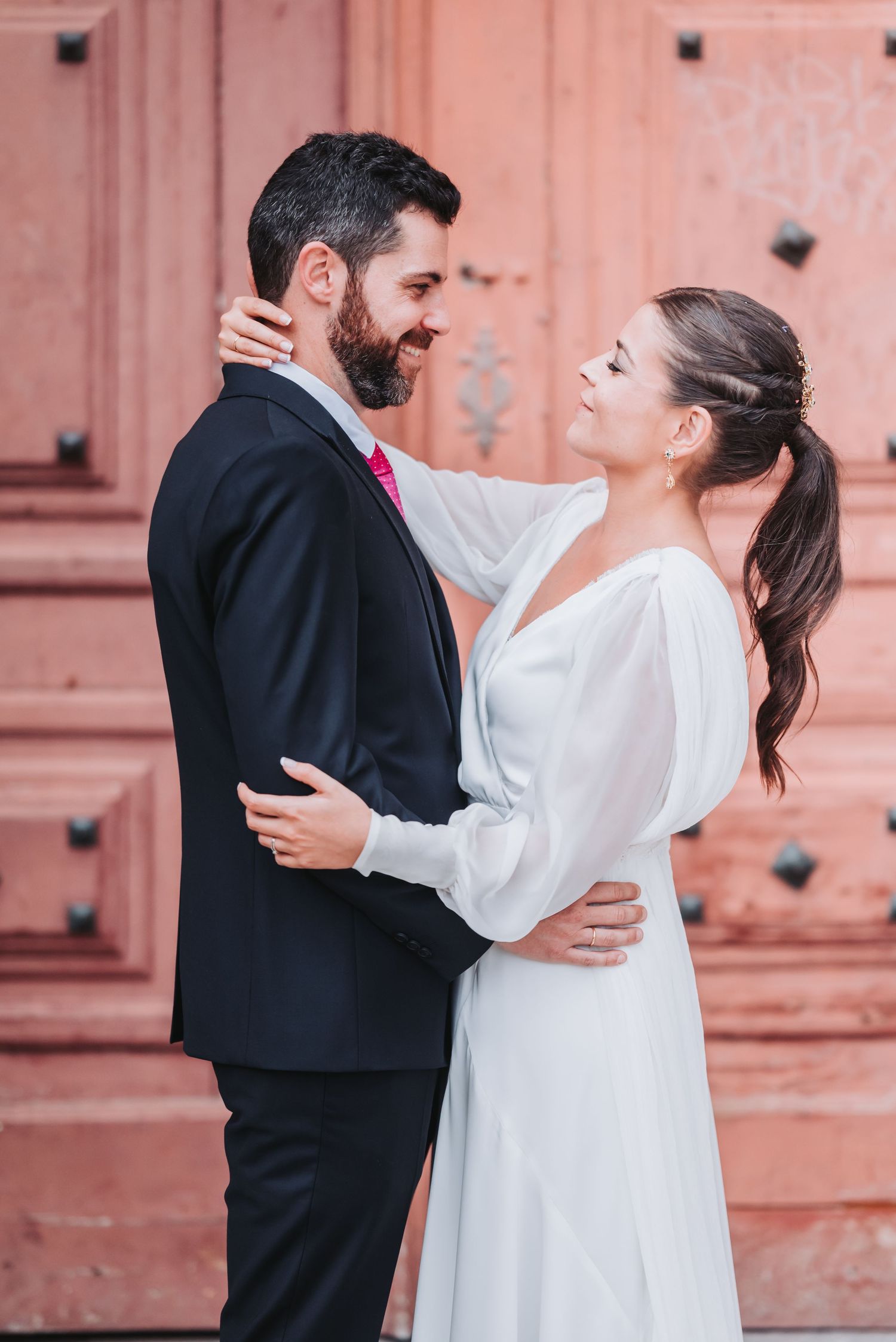 A romantic wedding couple embraces in front of a coral-colored door, with the bride in white and groom in black suit.
