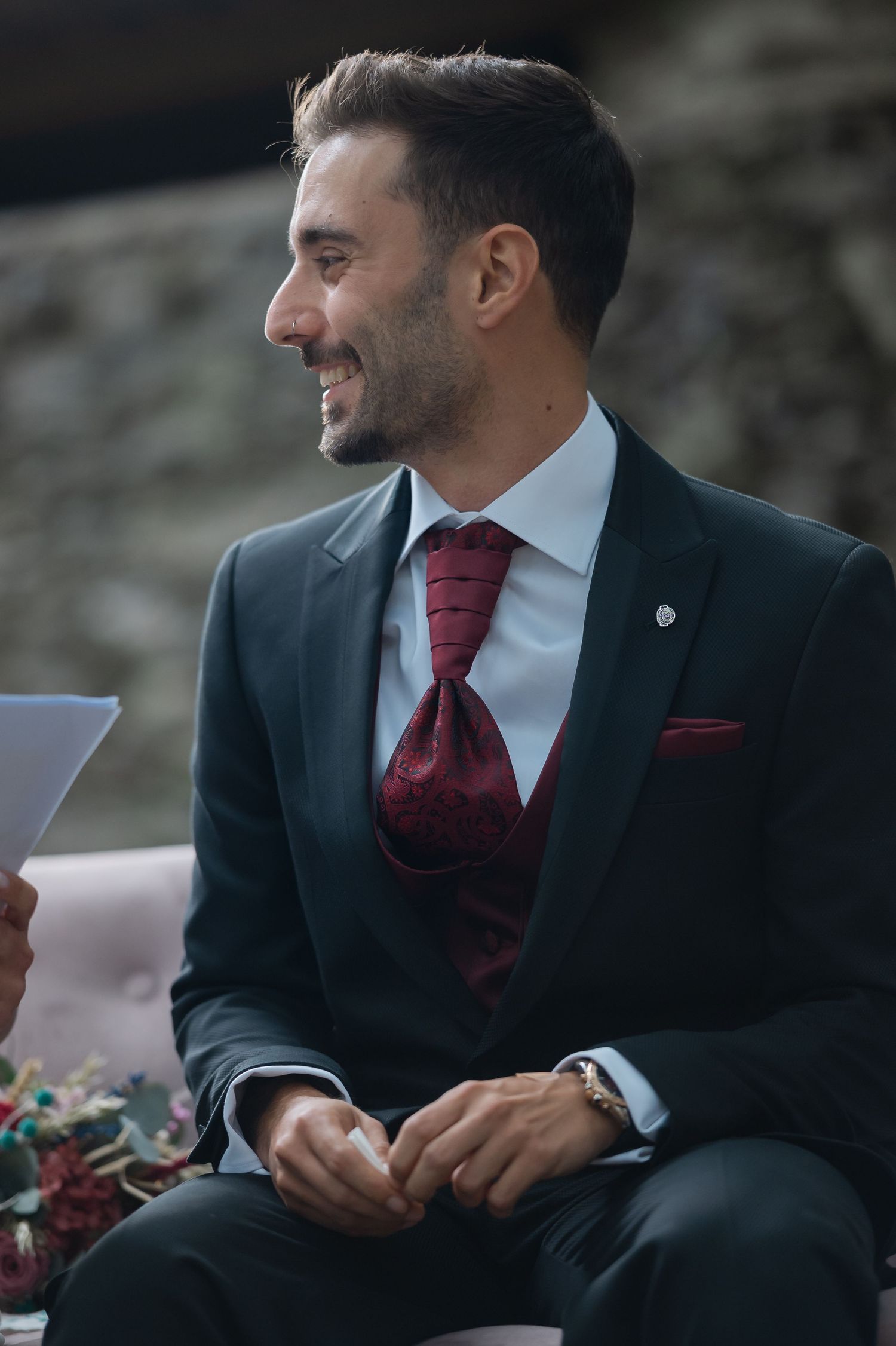 Groom in dark suit with burgundy tie smiles during outdoor wedding ceremony.